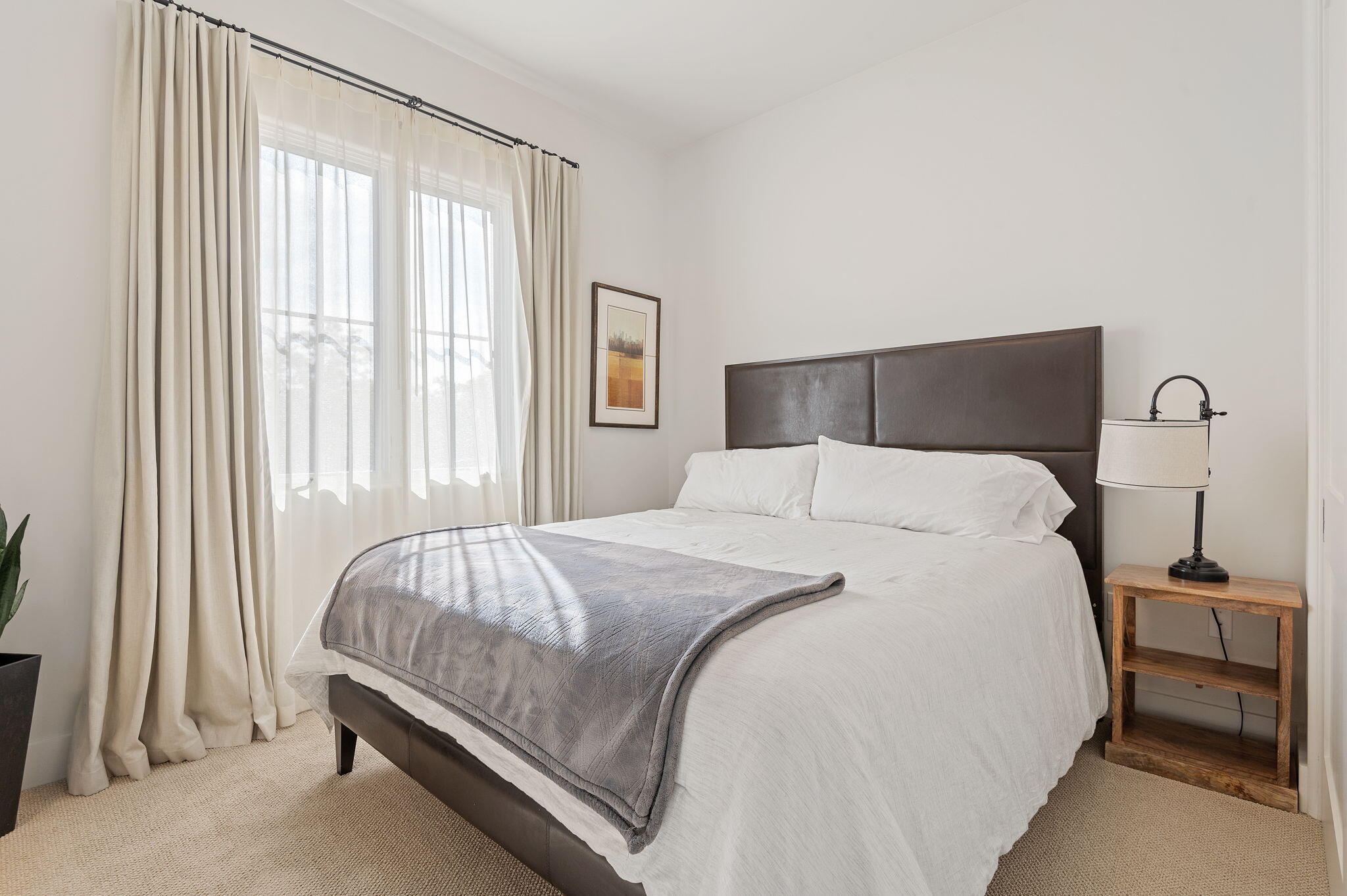 Bright bedroom with a large white bed, gray throw, dark headboard, and tall curtained windows.