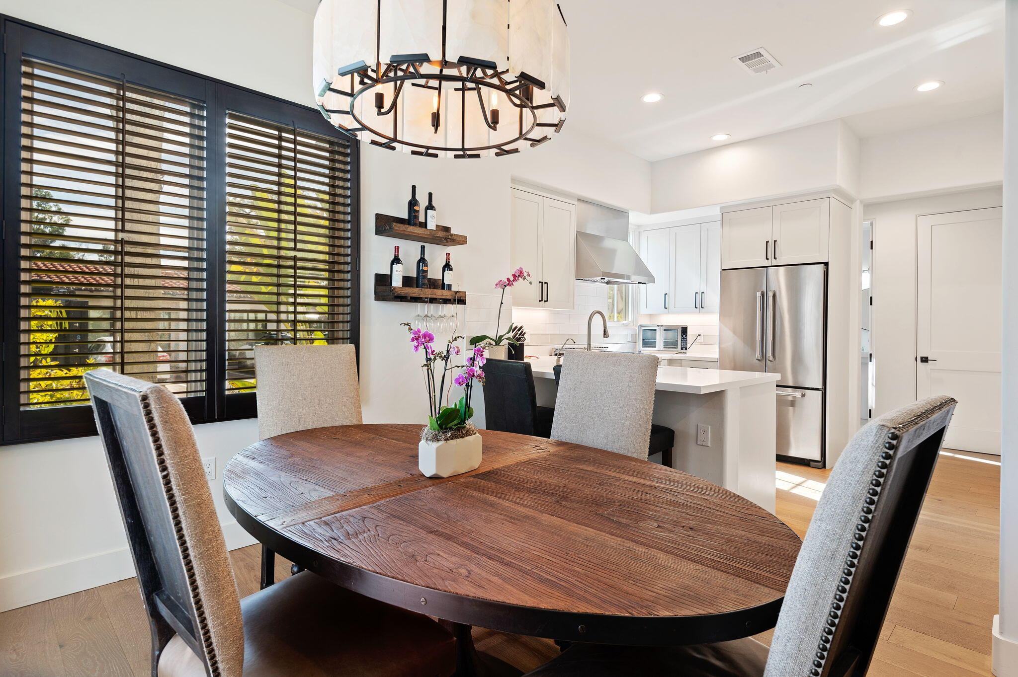 Dining area with a dark oval wood table beside a bright modern kitchen and wide shuttered windows.
