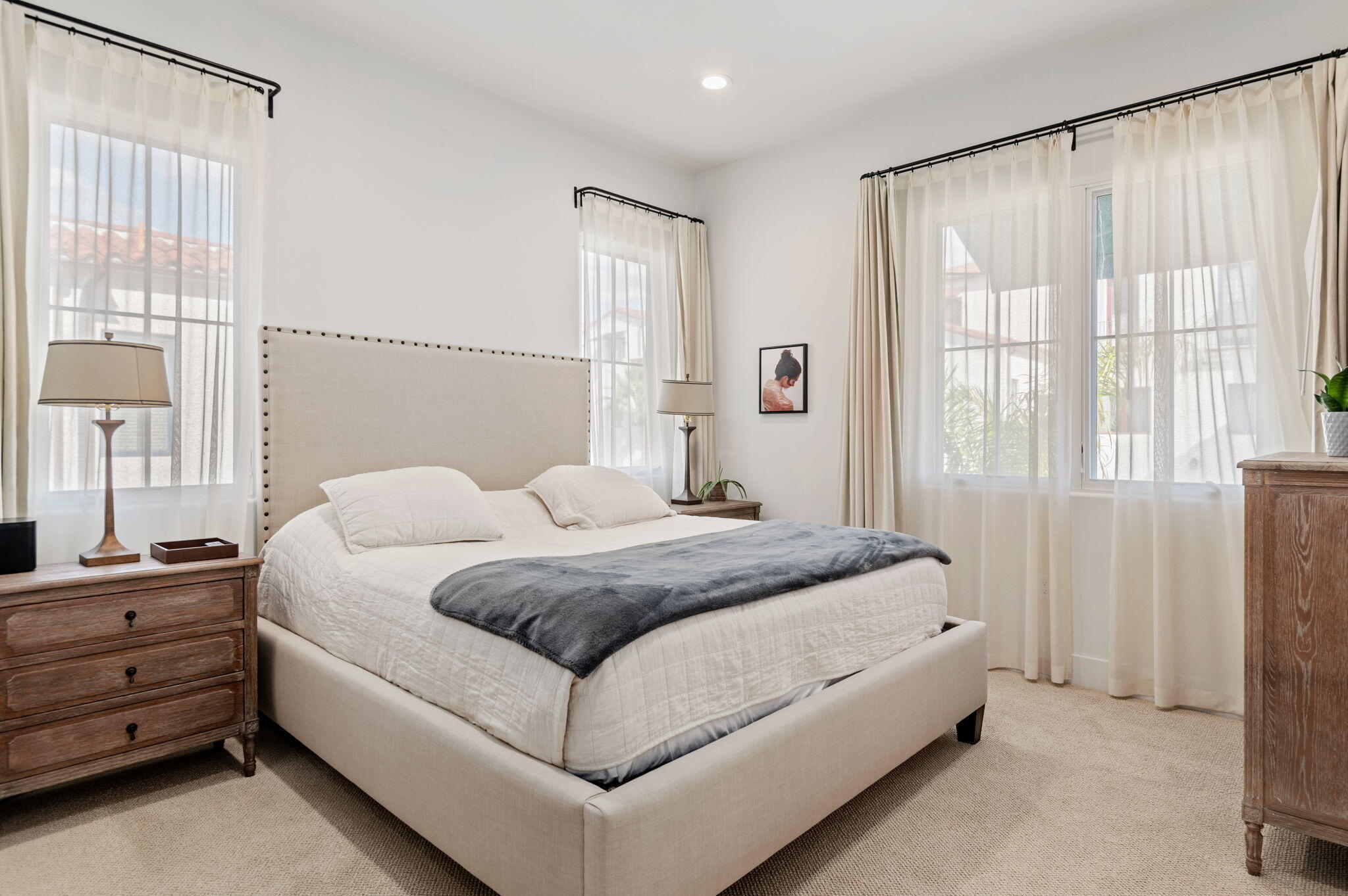 Bright bedroom with a large upholstered bed, gray throw, sheer curtains, and wooden dressers.