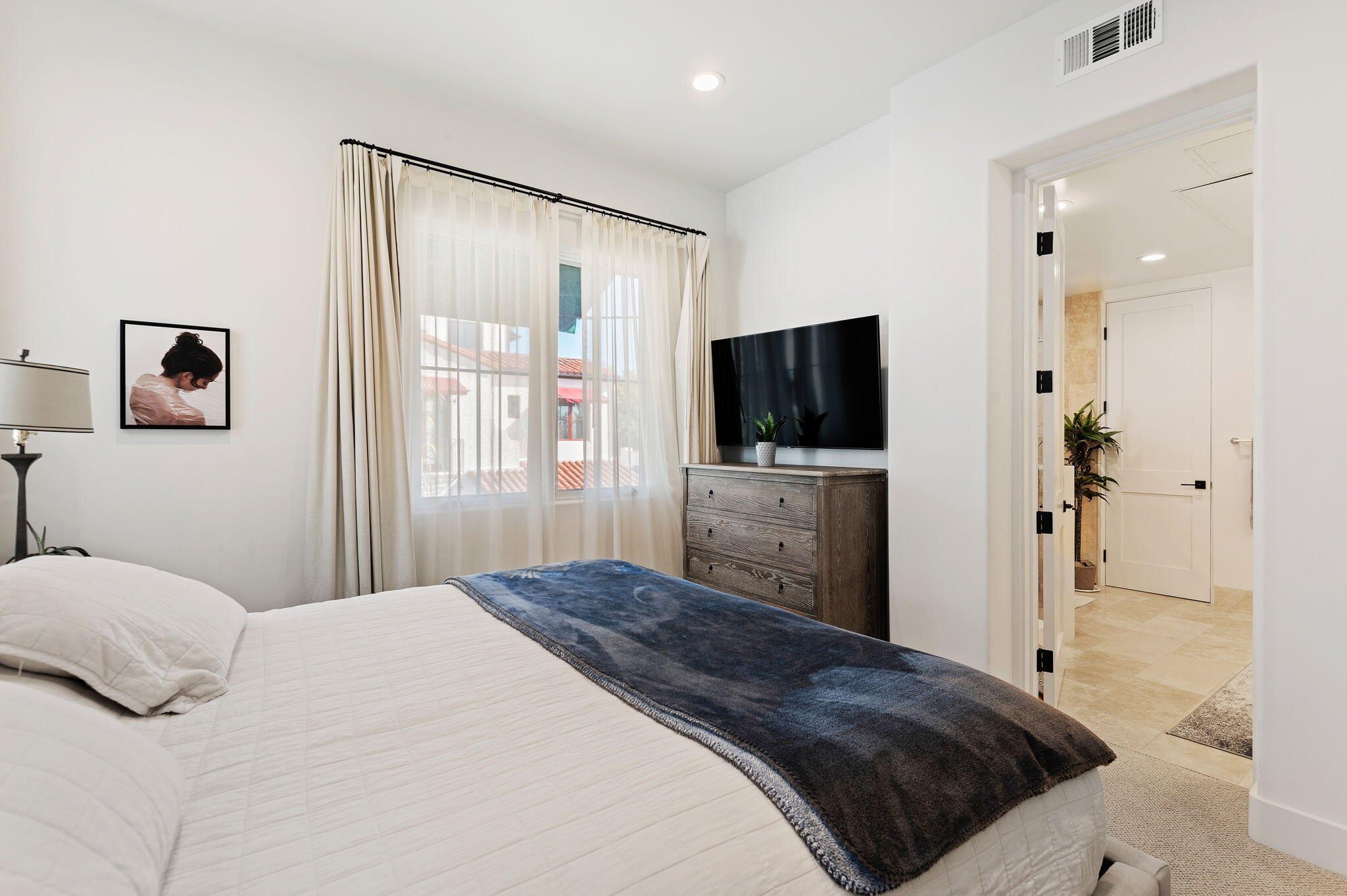 Bedroom with a white bed, dark blue throw, and television beside a curtained window.