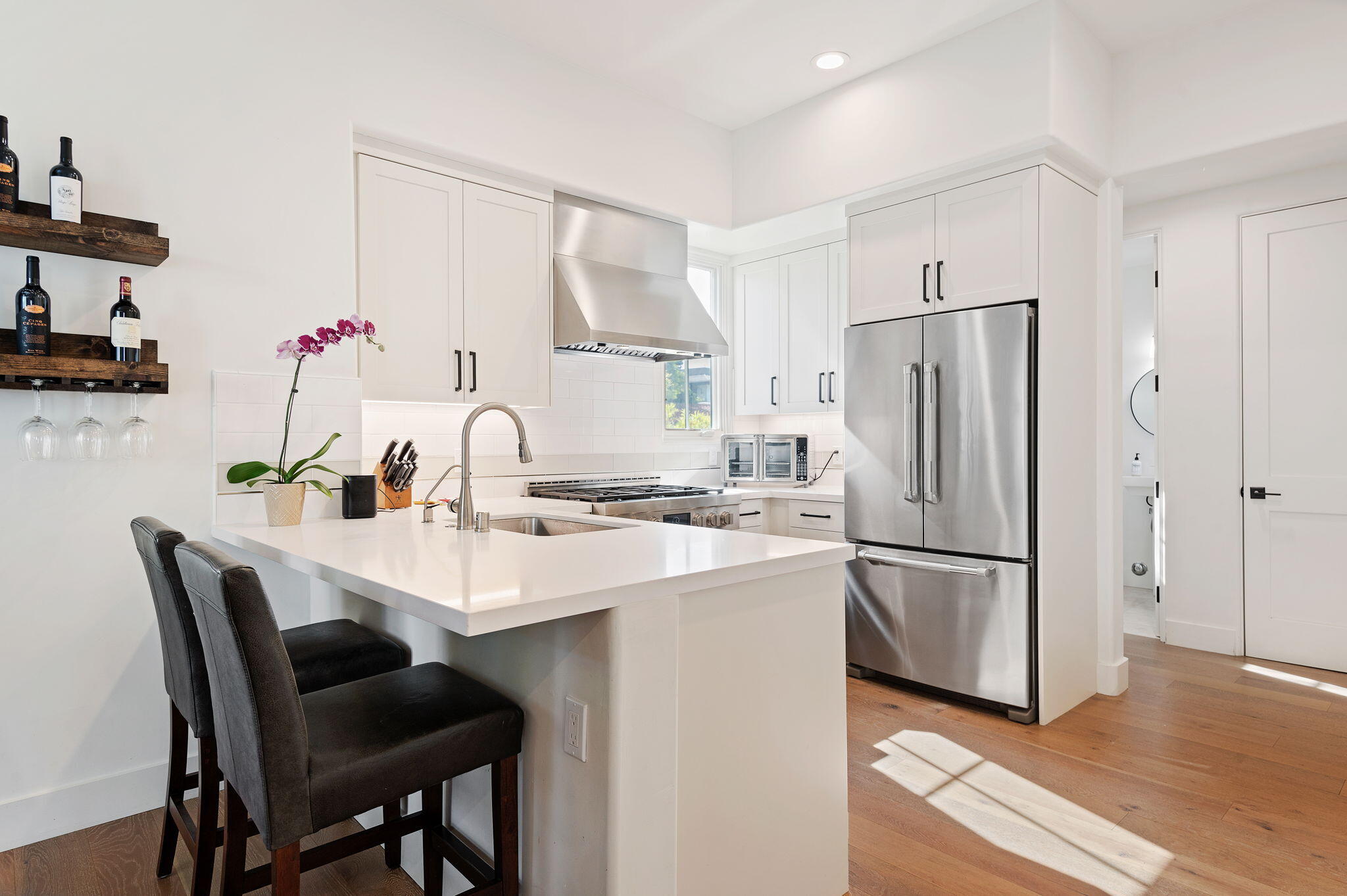 Modern white kitchen with a large island, stainless steel appliances, and floating wine shelves.