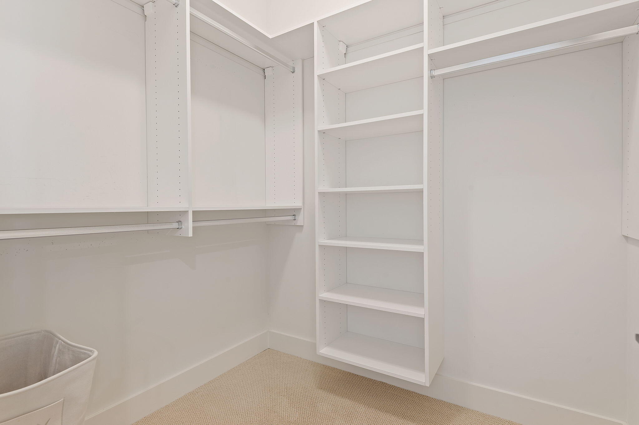 Walk-in closet with white built-in shelves, hanging rods, beige carpet, and a laundry hamper.