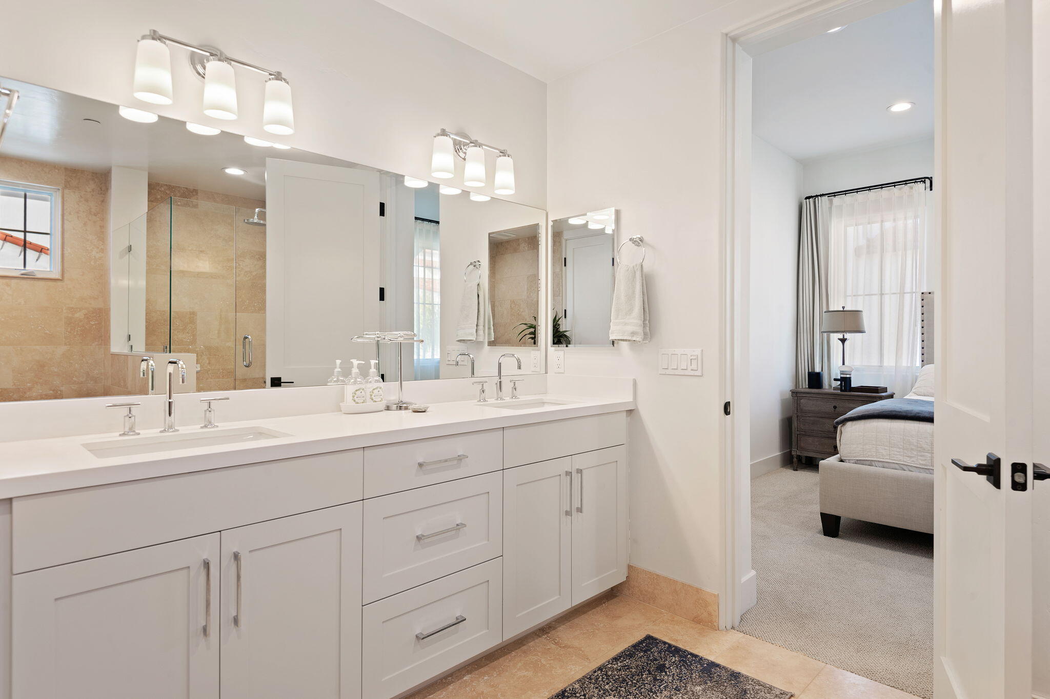 Bathroom with a white double vanity and tan tile, opening onto a furnished bedroom.