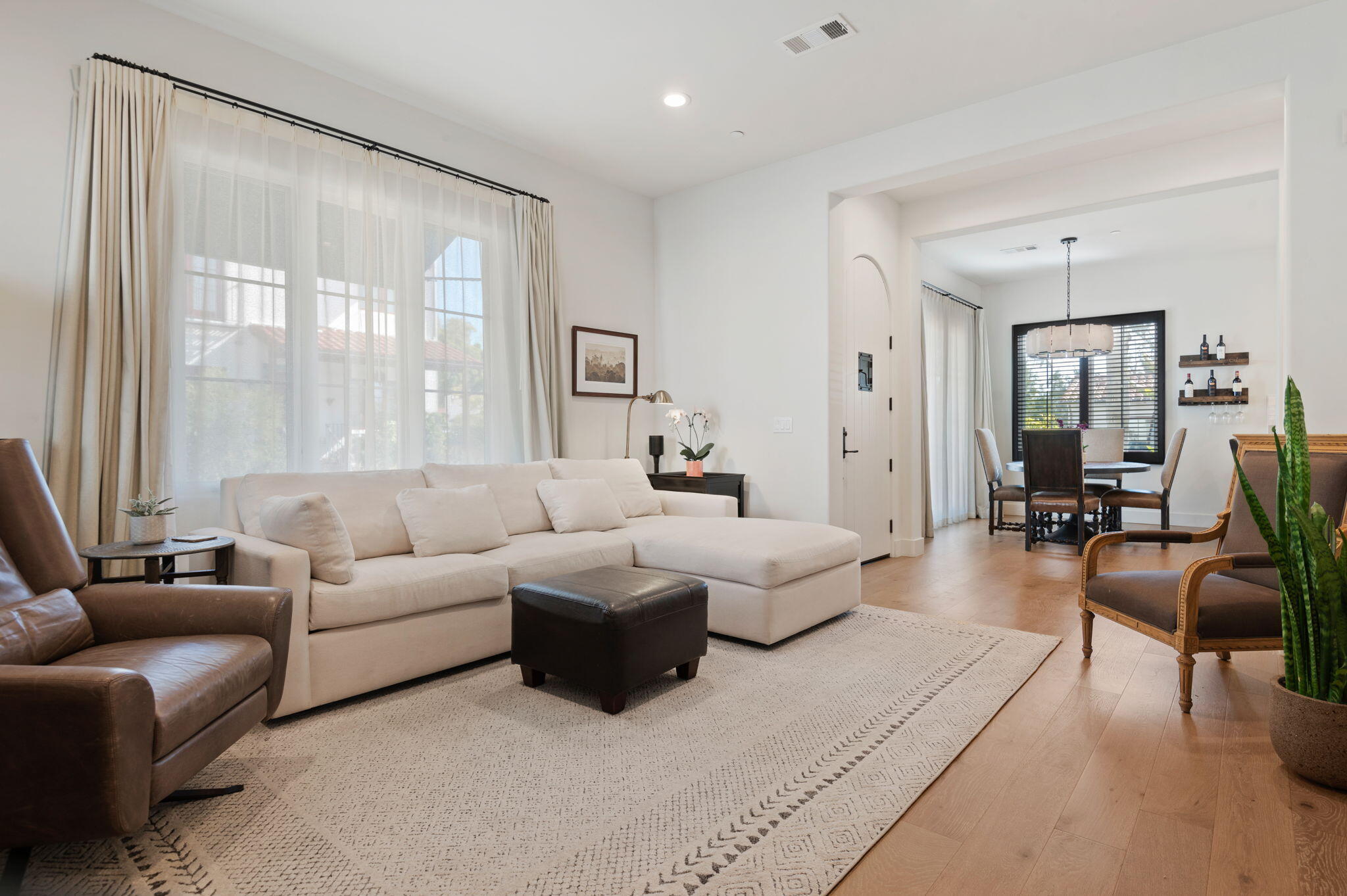 Furnished living room with a cream sectional, tall curtained windows, and an adjoining dining area.