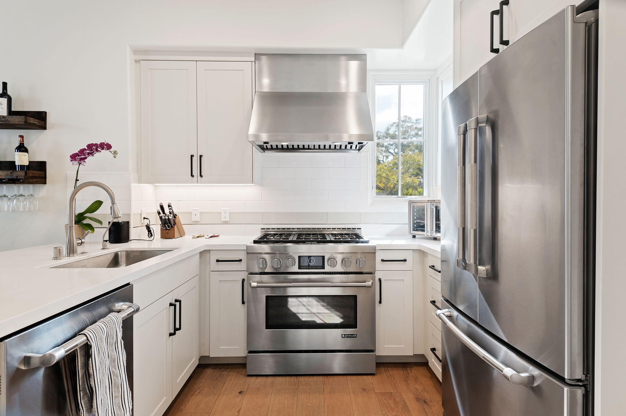 Bright white kitchen with stainless steel appliances, a large range hood, and warm wood floors.
