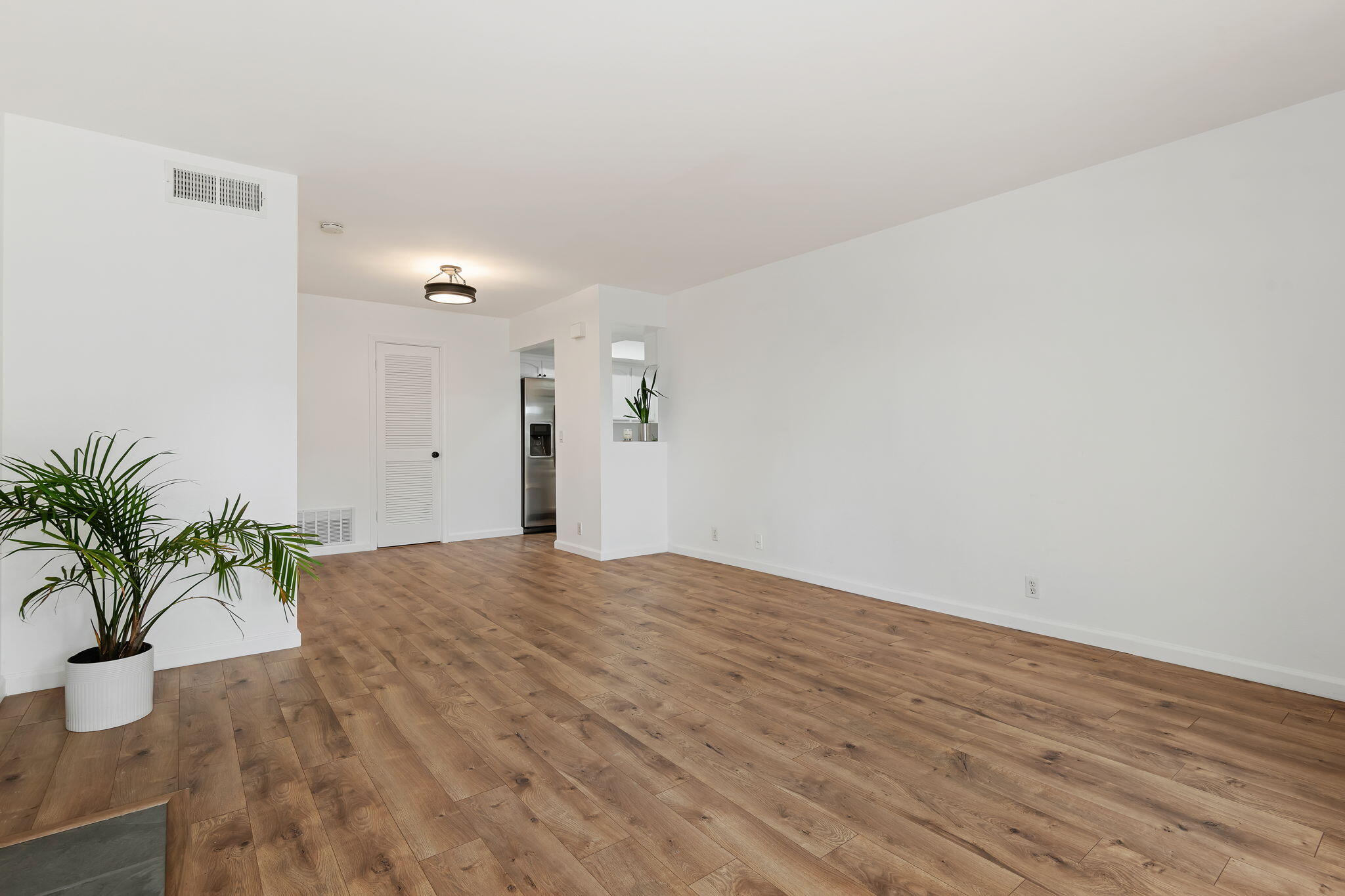 Empty living room with wide-plank wood floors, a potted palm, and a view into the kitchen.