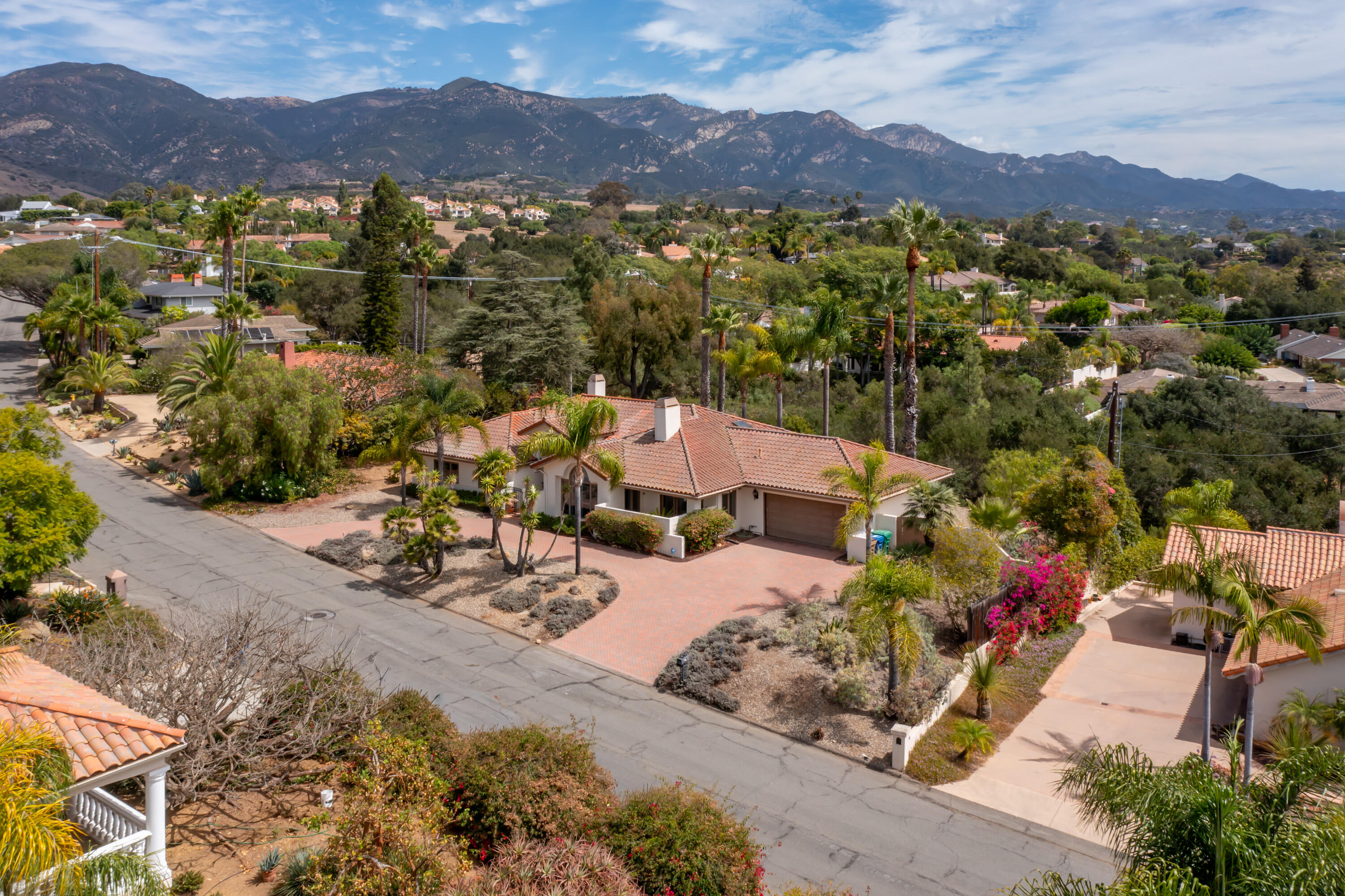Aerial view of a tile-roofed home with a brick driveway, palms, and mountains beyond.