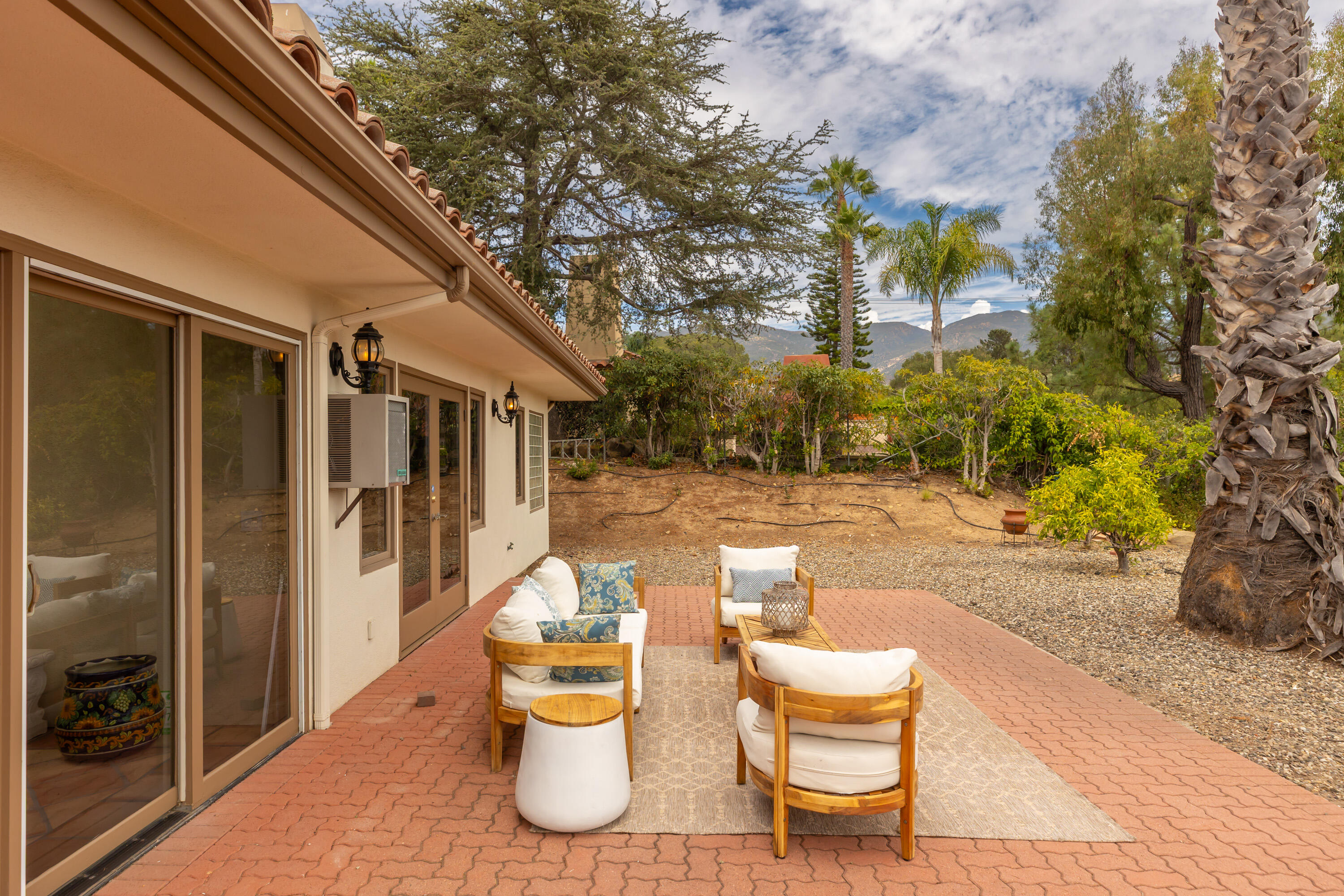 Furnished brick patio beside a home, with cushioned chairs, a dry garden, and mountains in the distance.