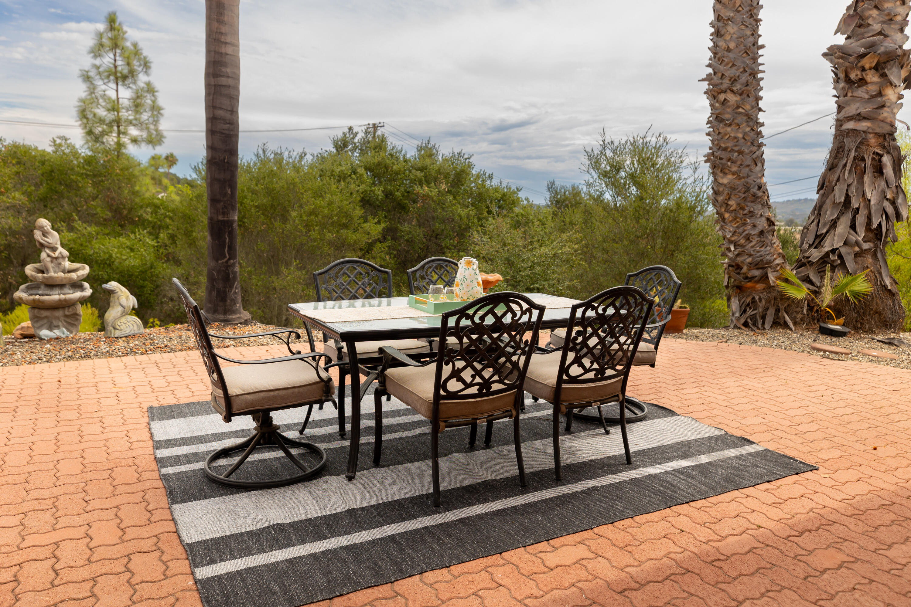 Outdoor dining patio with black metal furniture, striped rug, palm trees, and a stone fountain.