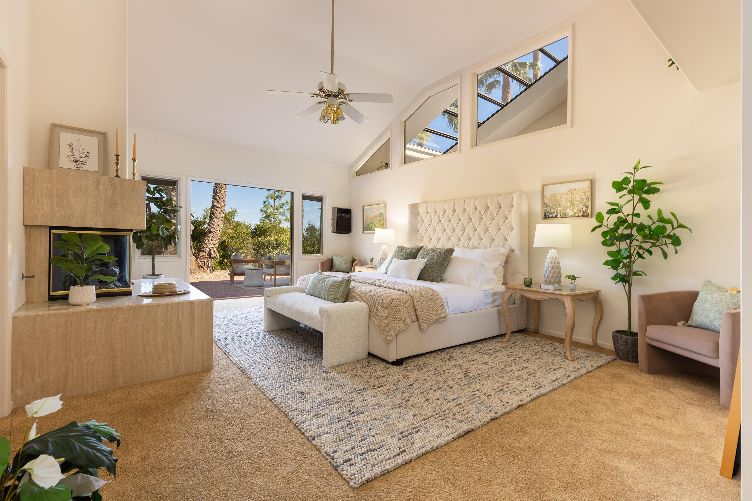 Bedroom with a tufted bed beside a stone fireplace and tall windows overlooking a leafy patio.