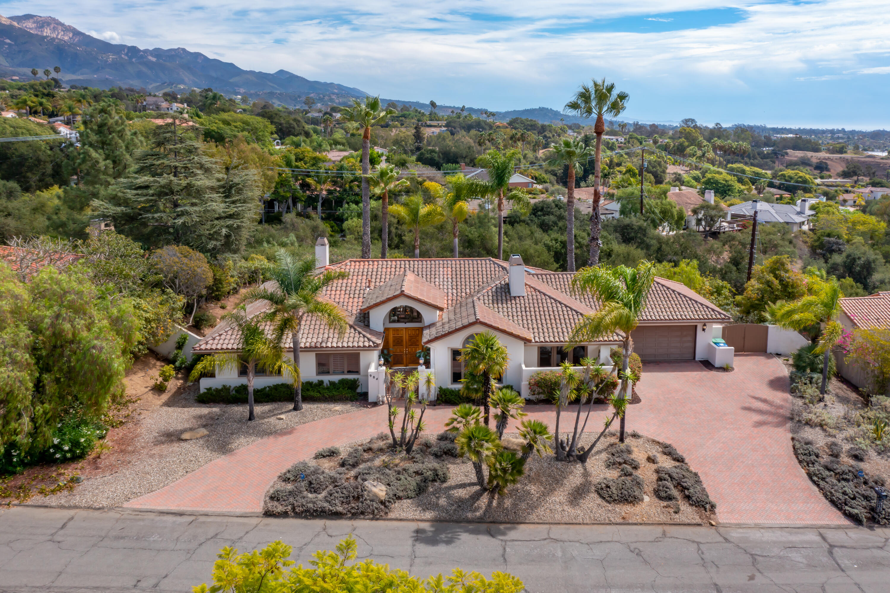White tile-roofed home with palm trees and brick driveway, backed by green hills and mountains.