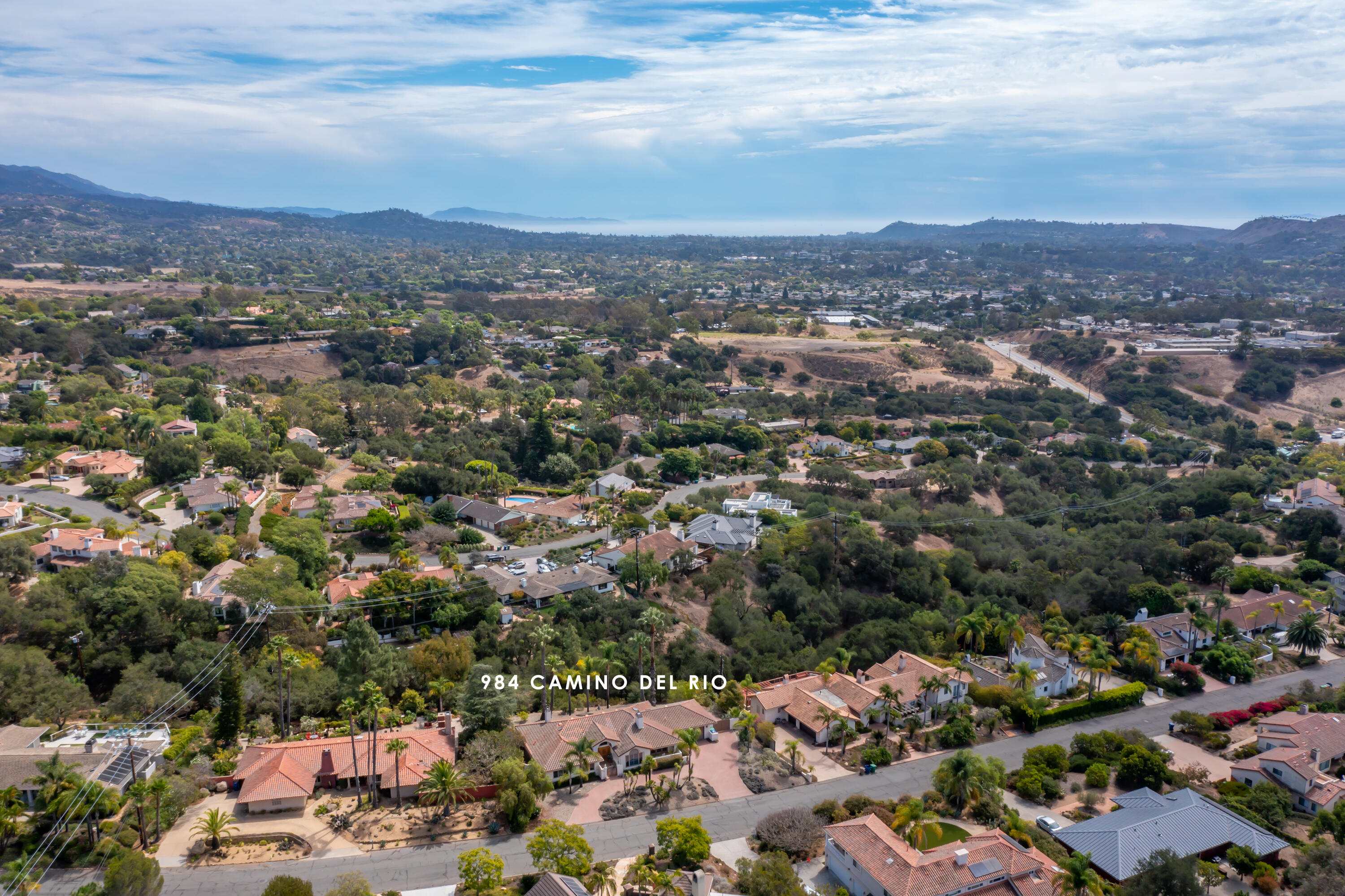 Aerial view of 984 Camino Del Rio amid tree-lined homes, with hills and distant ocean.