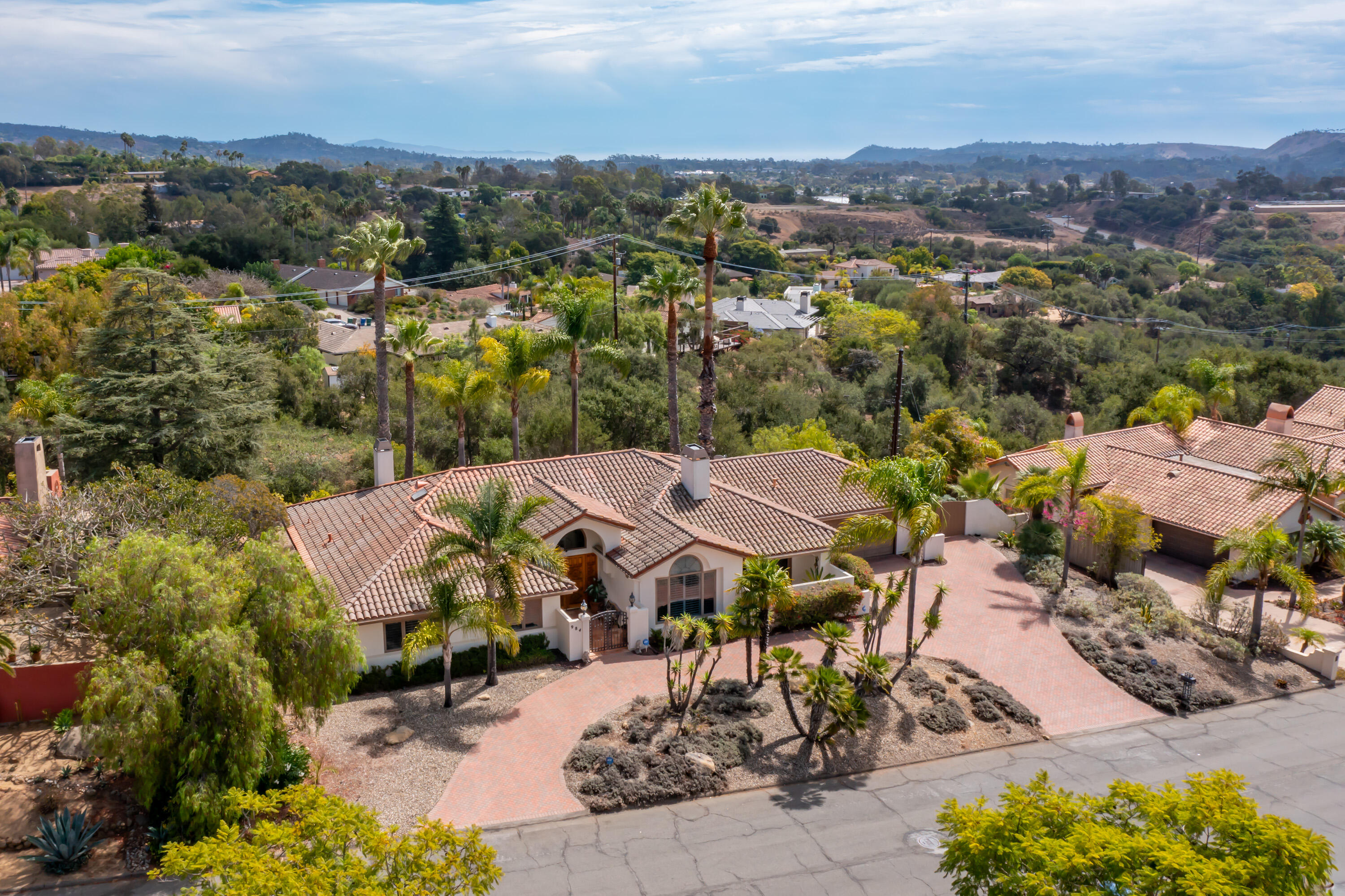 Aerial view of a tile-roofed home with a brick driveway, palm trees, and distant hills.