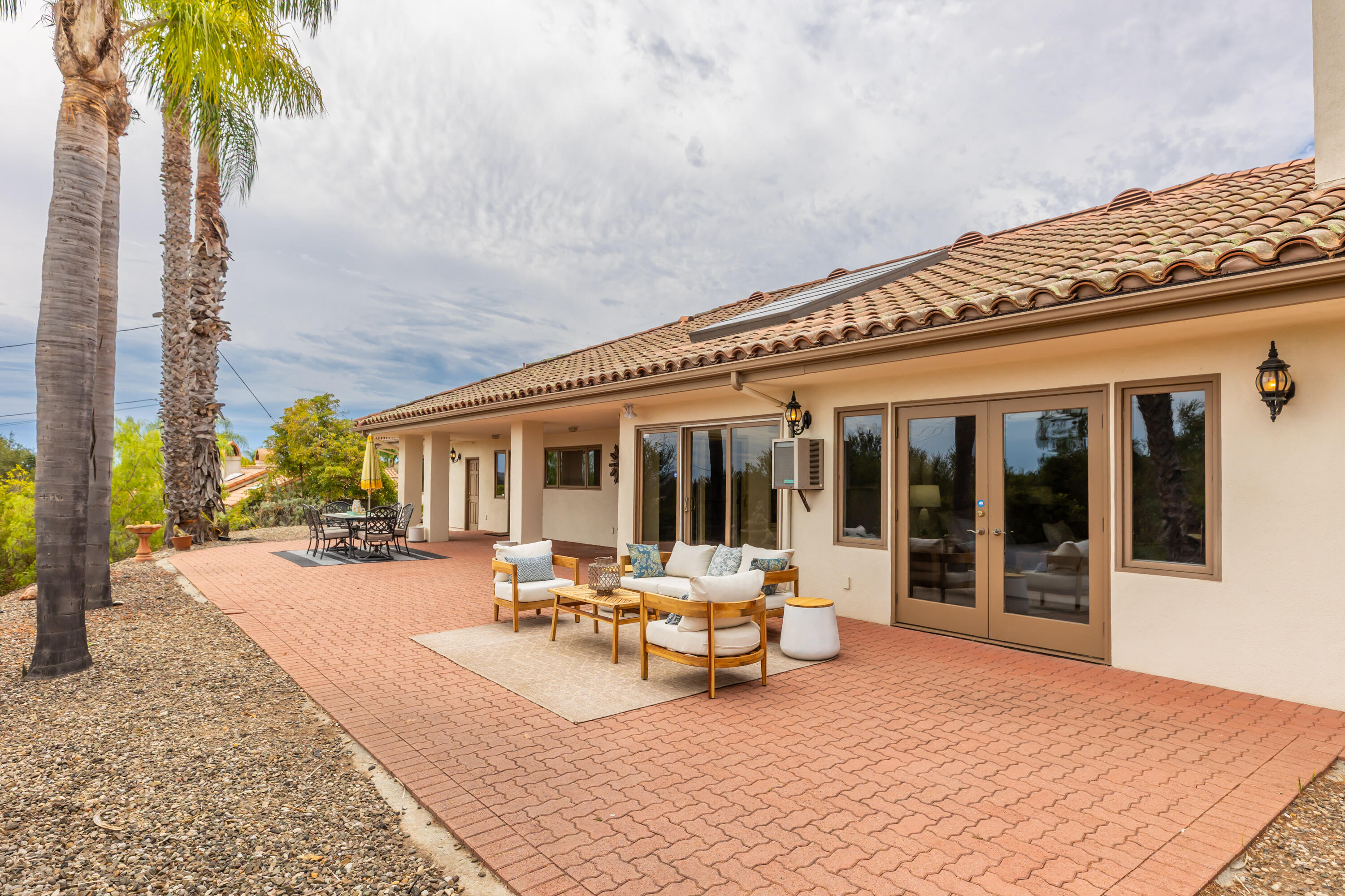 Home’s rear patio with brick paving, outdoor lounge seating, dining table, and tall palm trees.