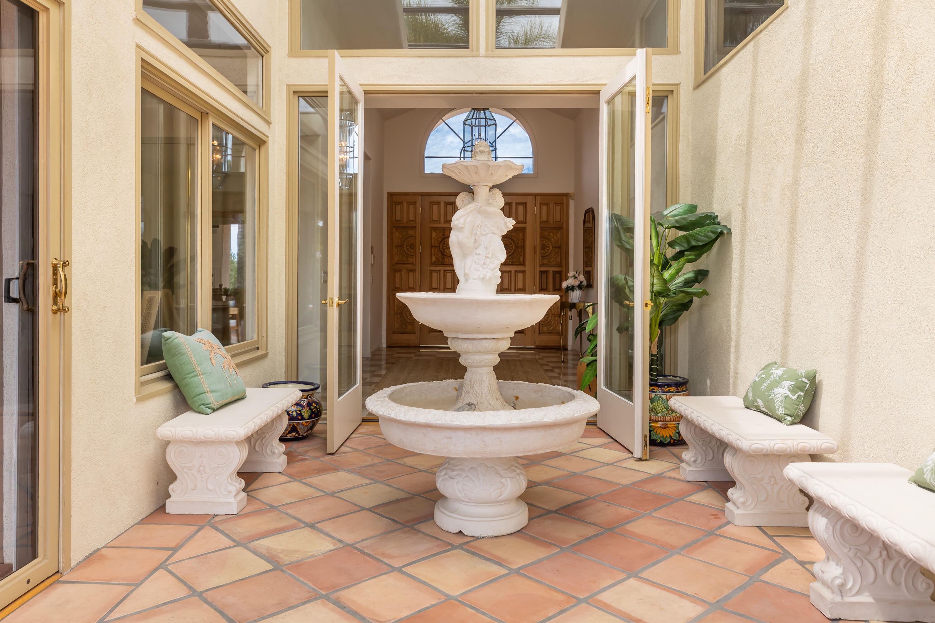 Tiled entry courtyard with an ornate white fountain, stone benches, and open glass doors.