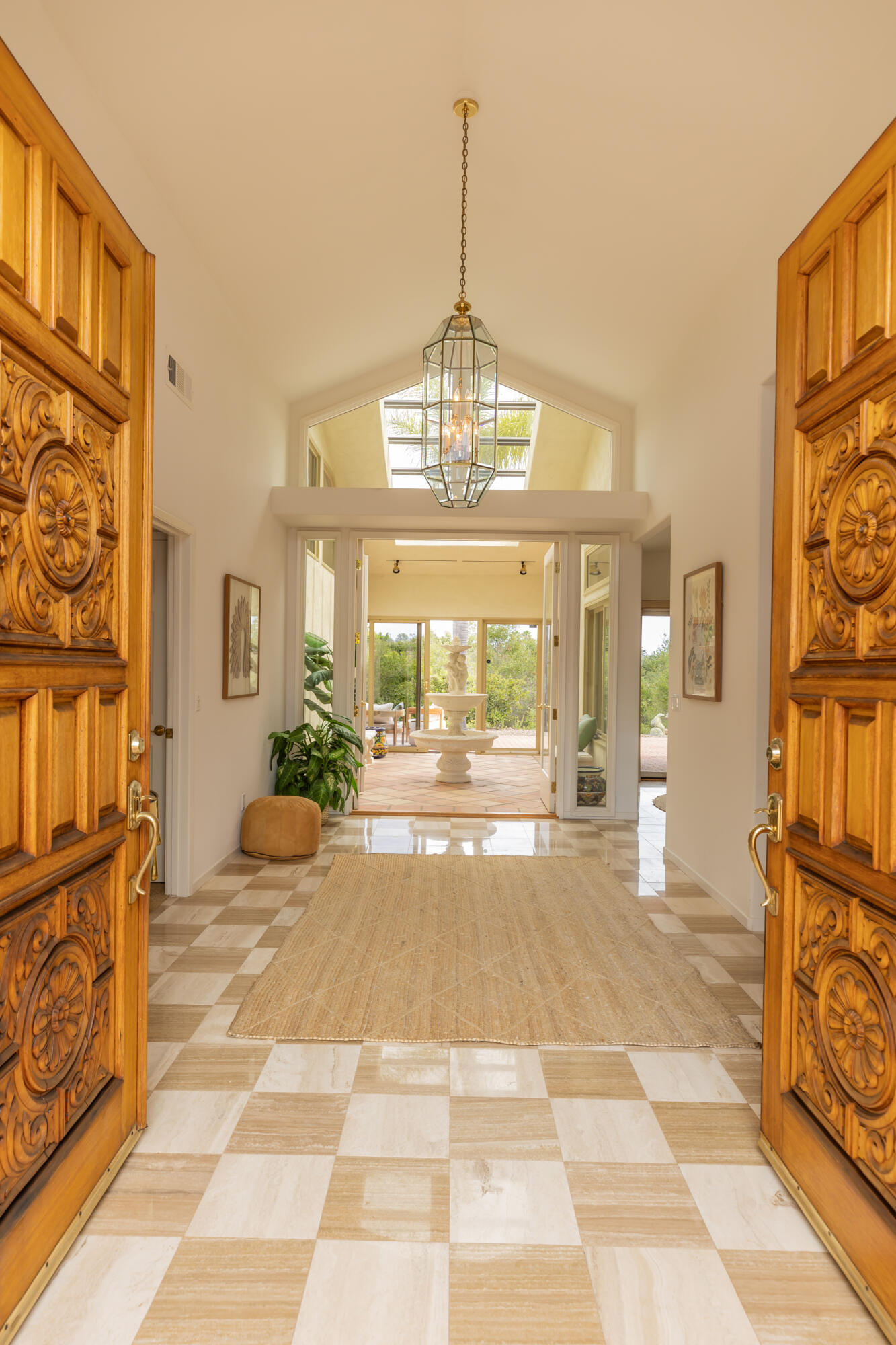 Entry foyer with carved wooden doors, a glass chandelier, checkerboard tile, and a courtyard fountain beyond.