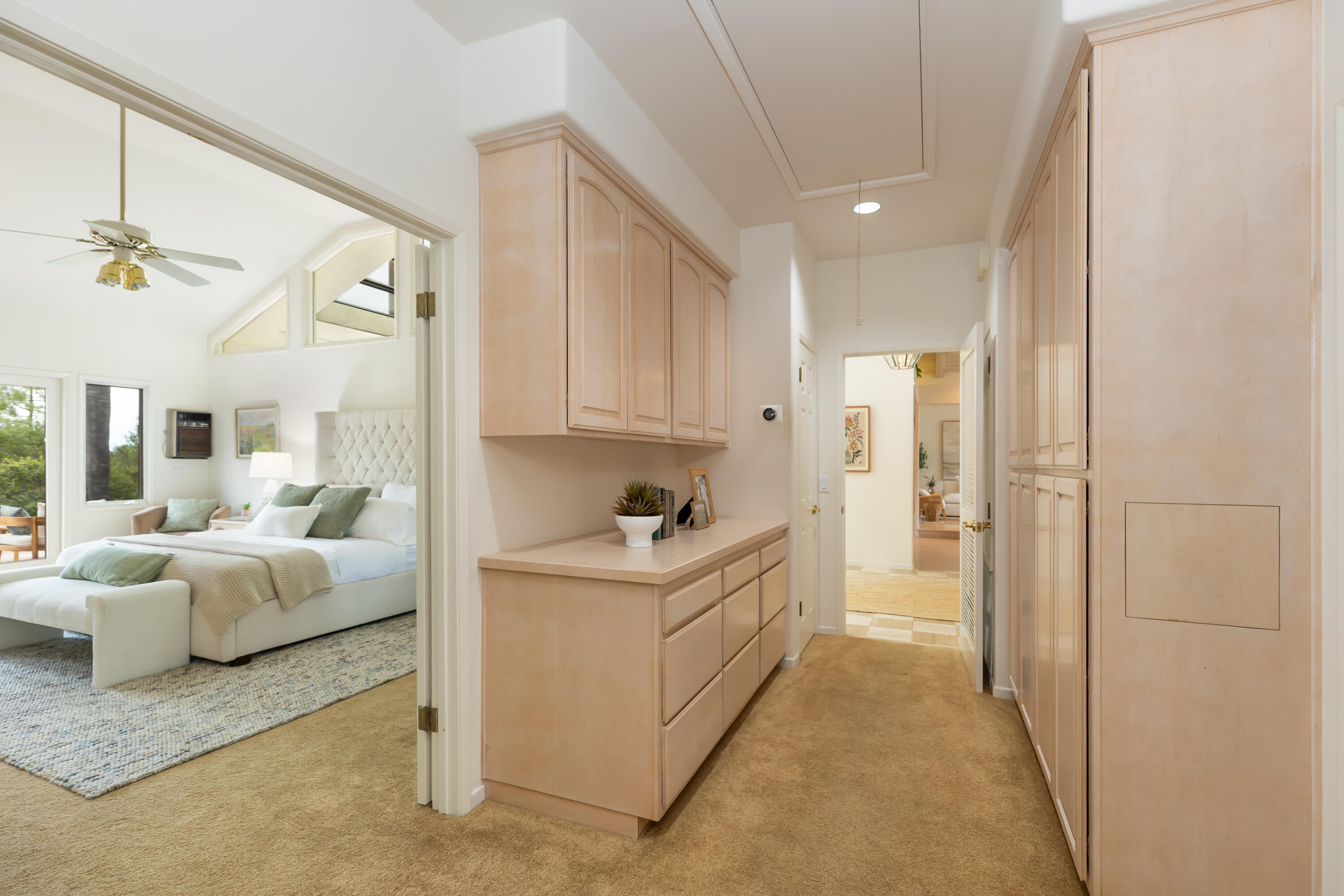 Dressing hallway with built-in light wood cabinets beside a bright bedroom with a tufted headboard.
