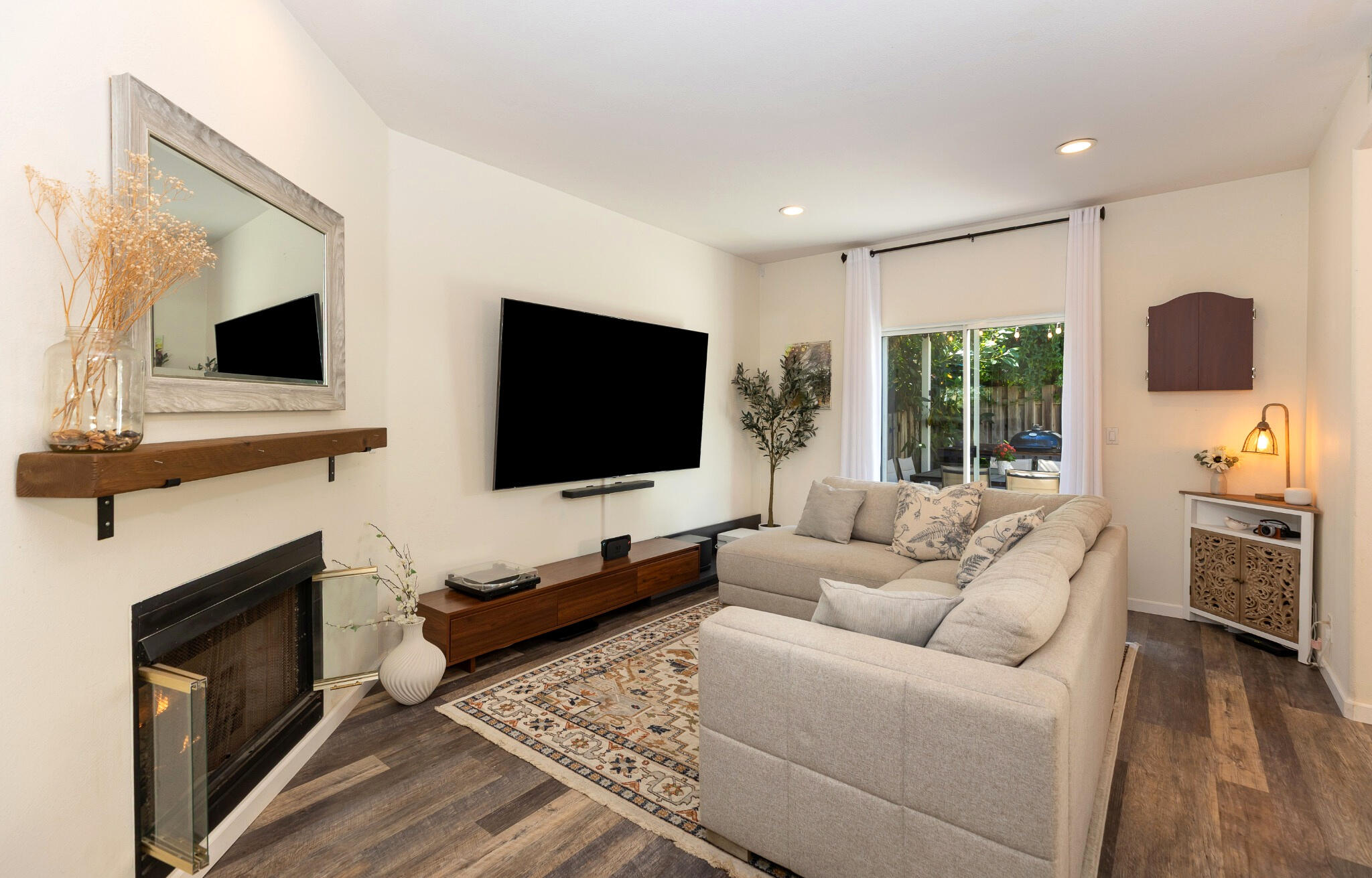 Living room with a sectional sofa, fireplace, wall-mounted television, and sliding glass doors to a leafy patio.