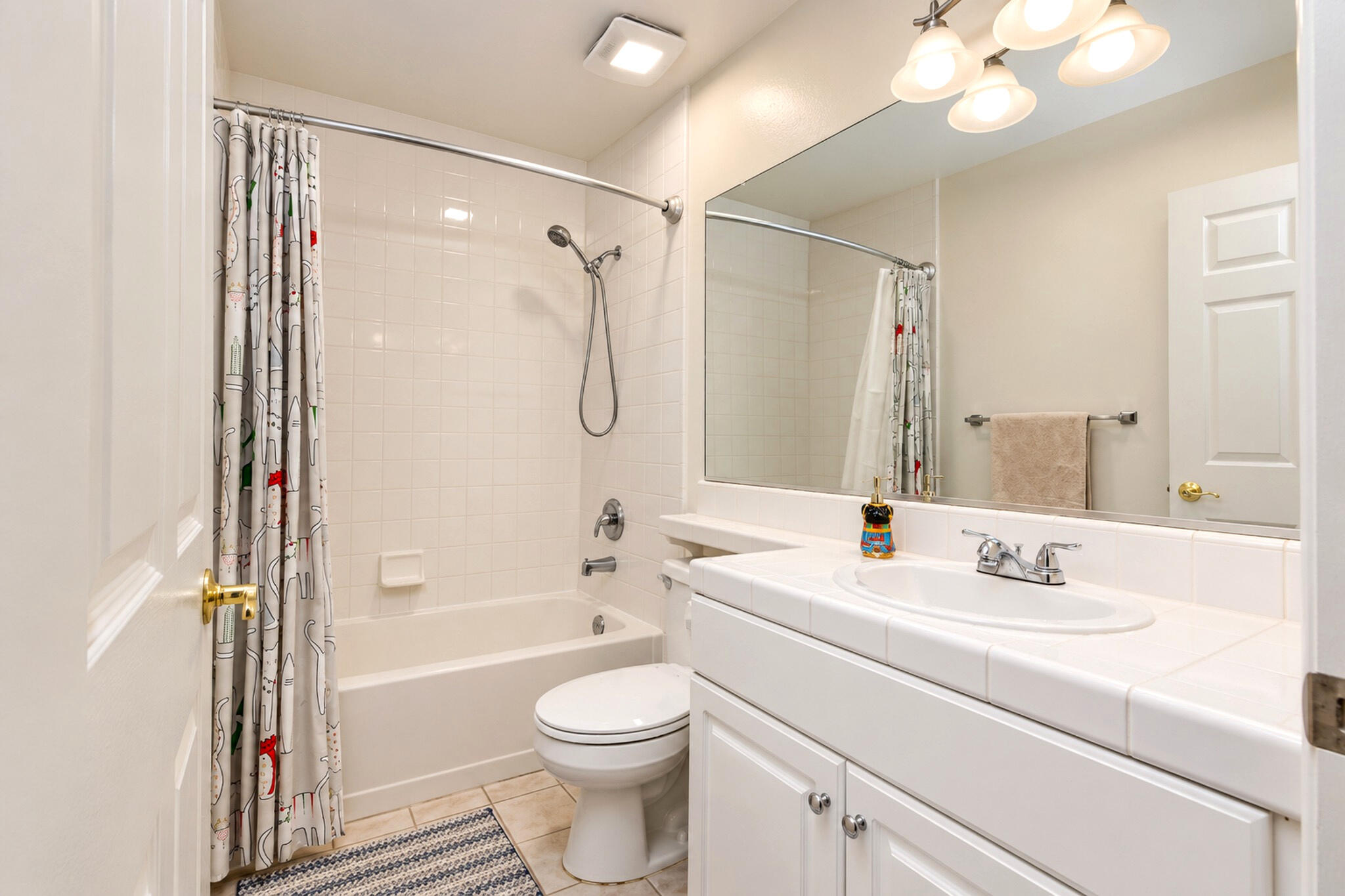 Bathroom with a white bathtub, patterned shower curtain, toilet, and tiled vanity beneath a large mirror.