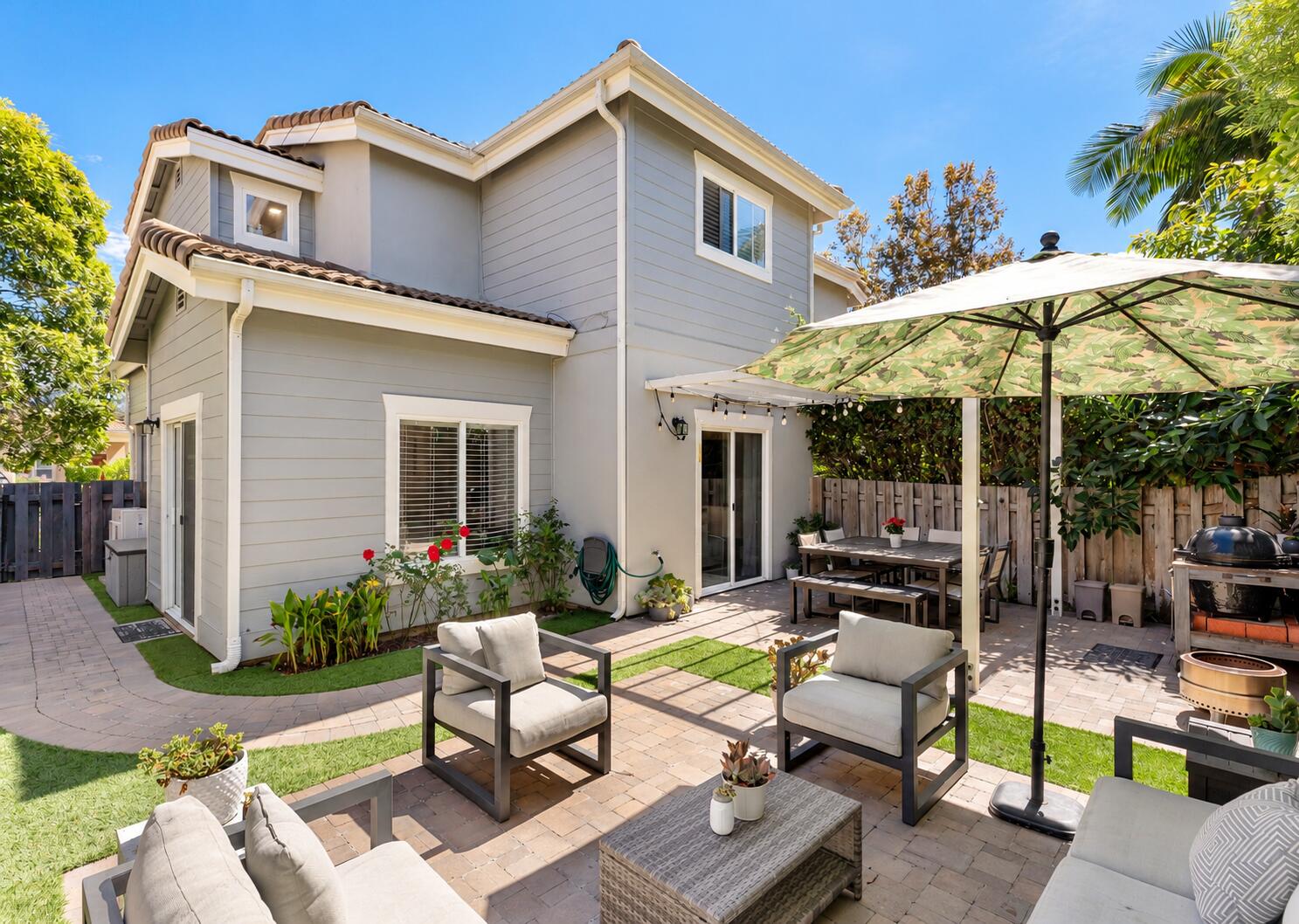Two-story gray house with a furnished backyard patio, dining area, patterned umbrella, and barbecue.