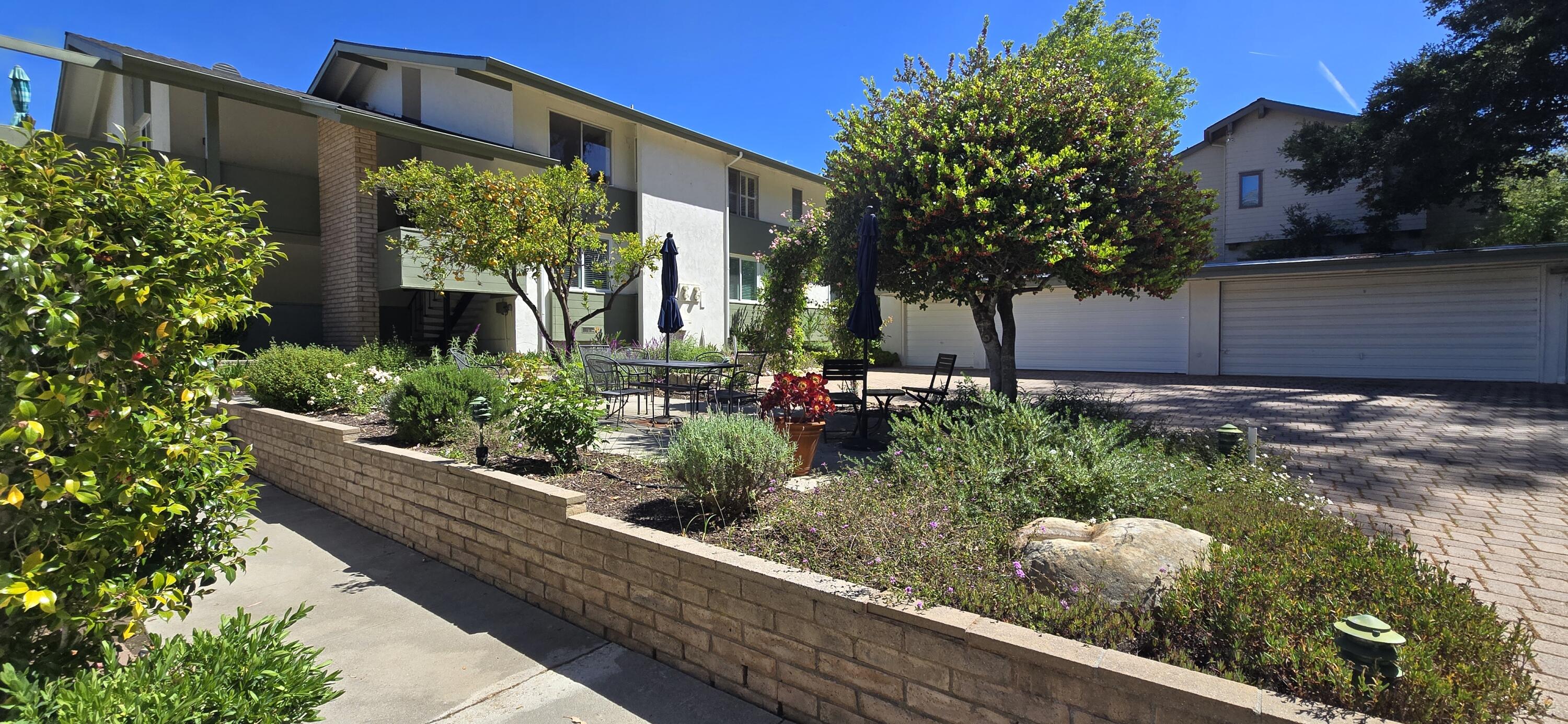 Apartment building courtyard with brick planters, mature trees, outdoor dining tables, and garages.