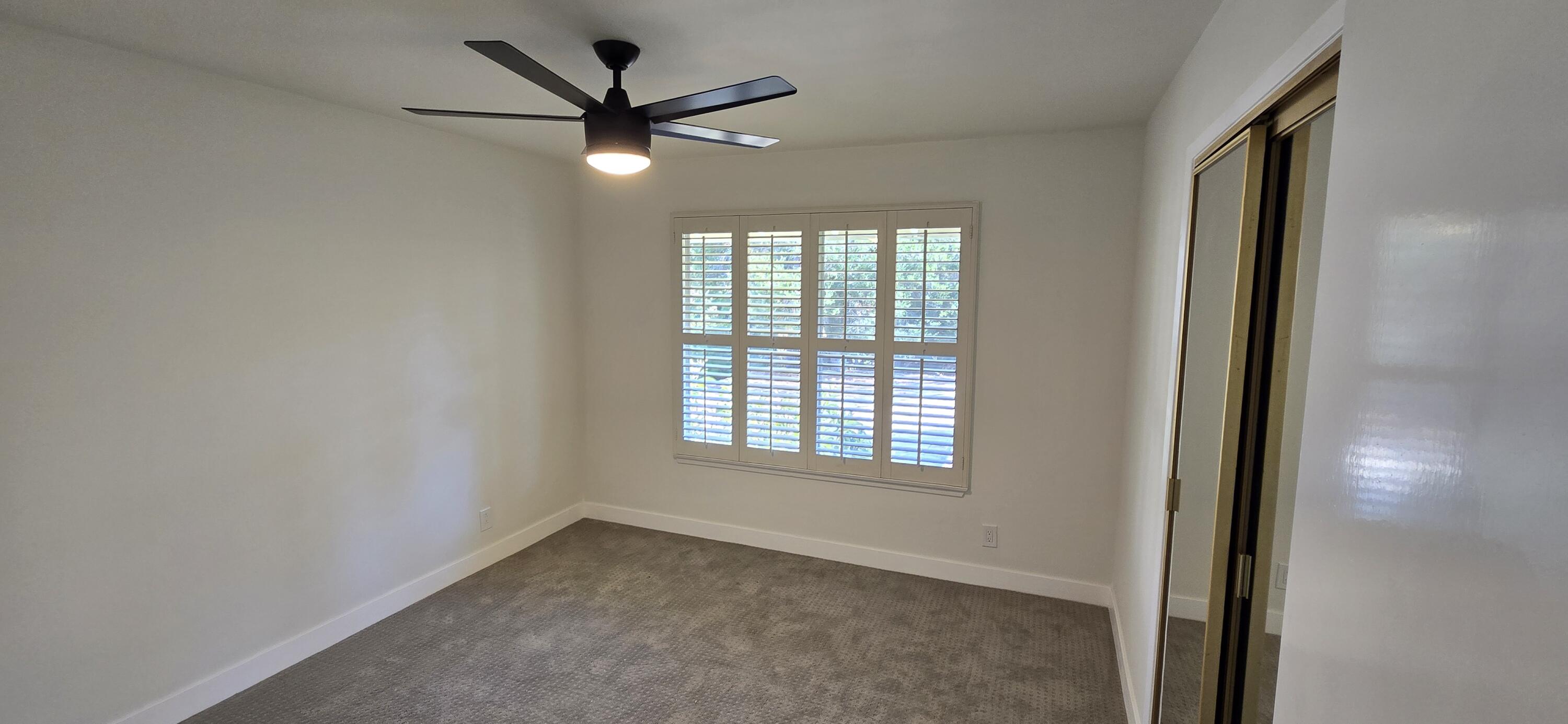 Empty carpeted bedroom with plantation shutters, a ceiling fan, and a mirrored closet door.