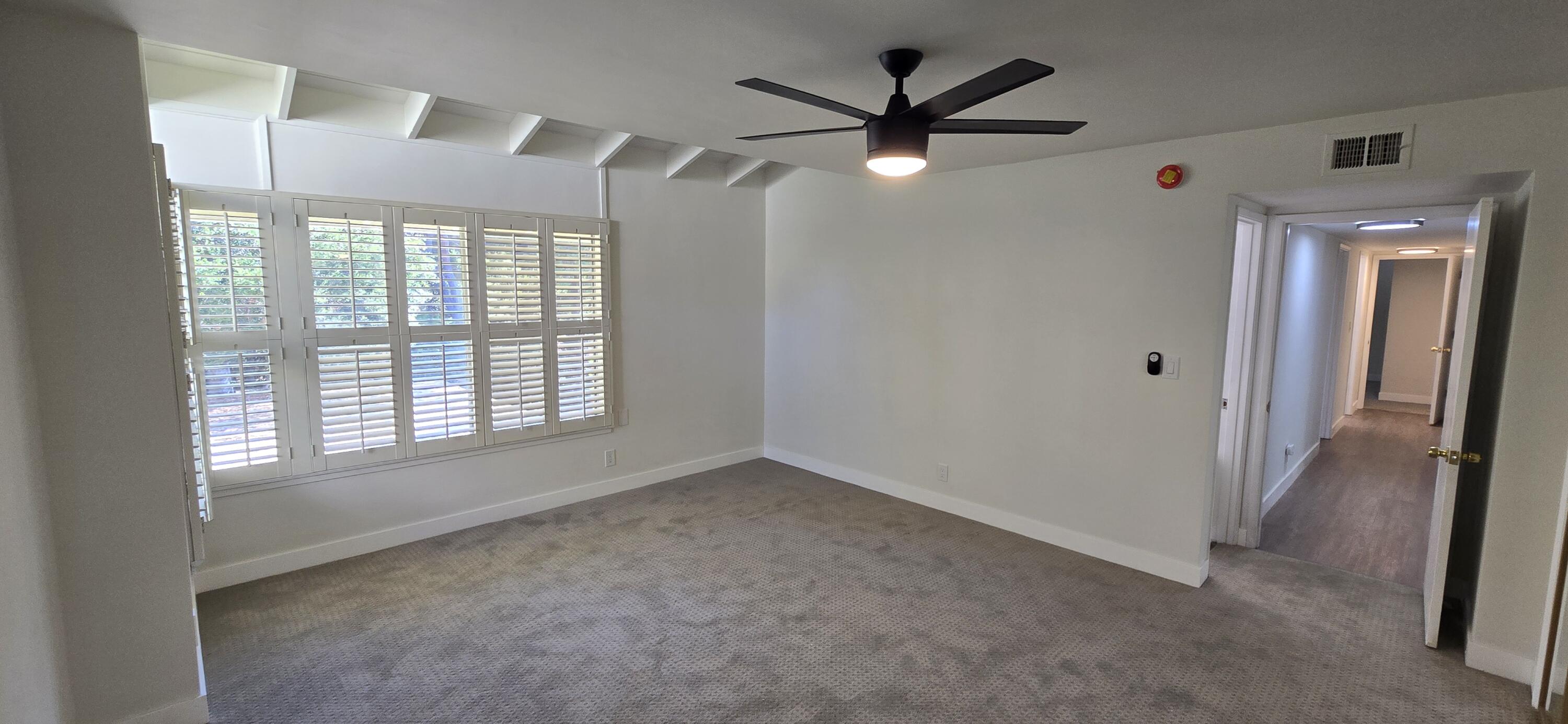 Vacant carpeted room with plantation shutters and a black ceiling fan, opening onto a hallway.