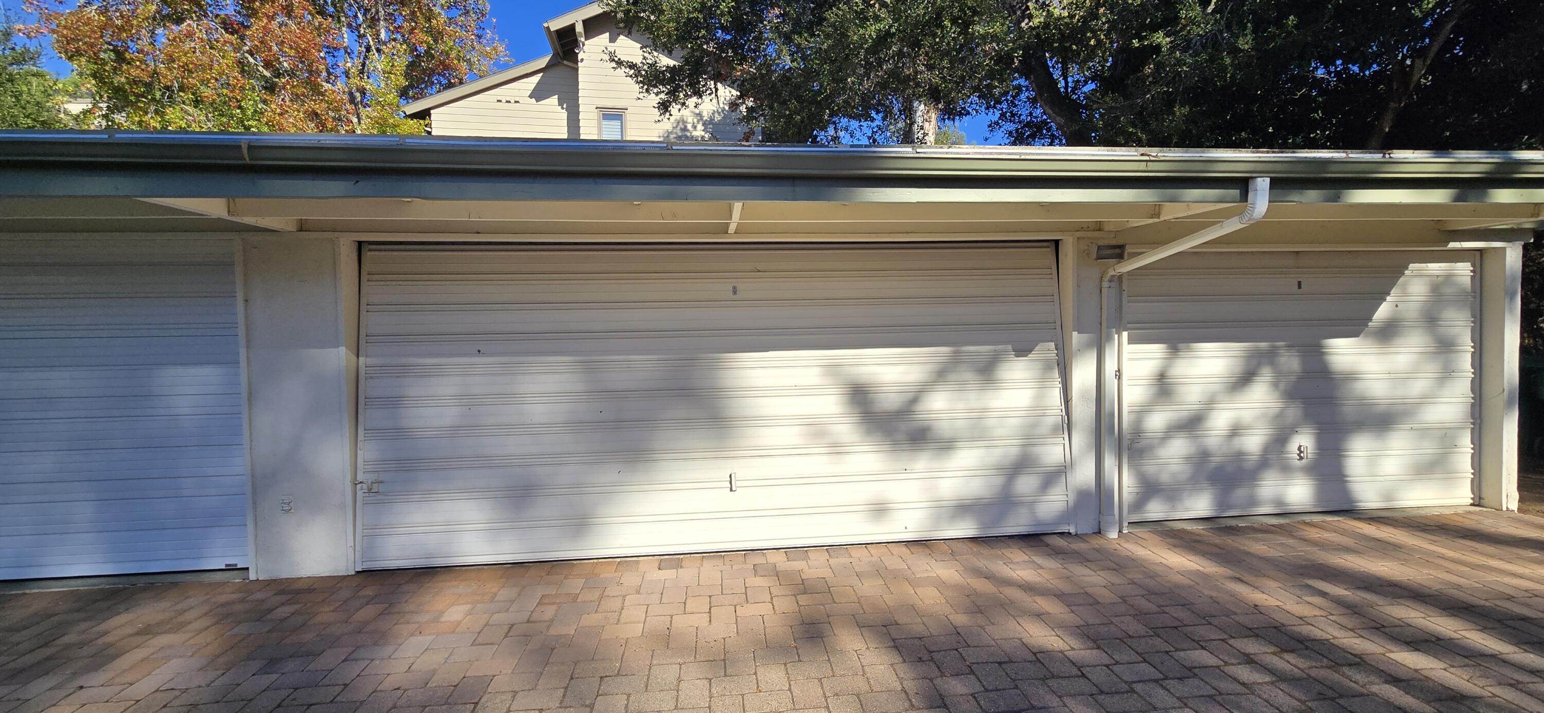 Three garage doors and a brick-paver driveway beneath mature trees.