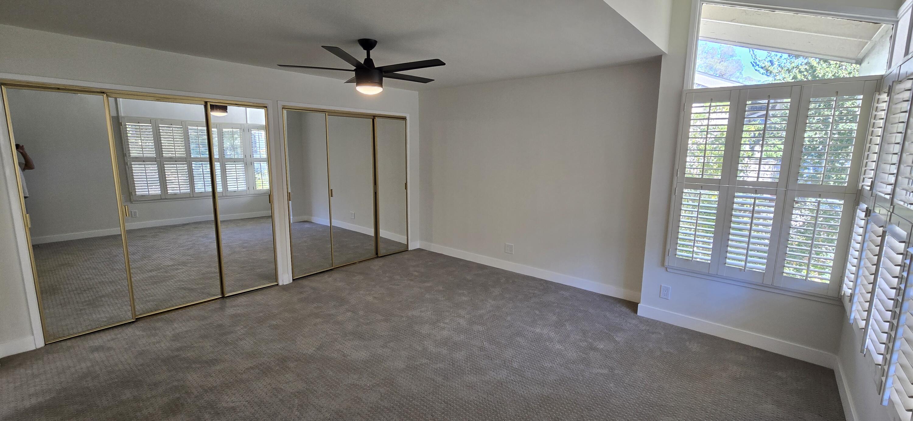 Unfurnished bedroom with mirrored closet doors, gray carpet, a ceiling fan, and plantation-shuttered windows.