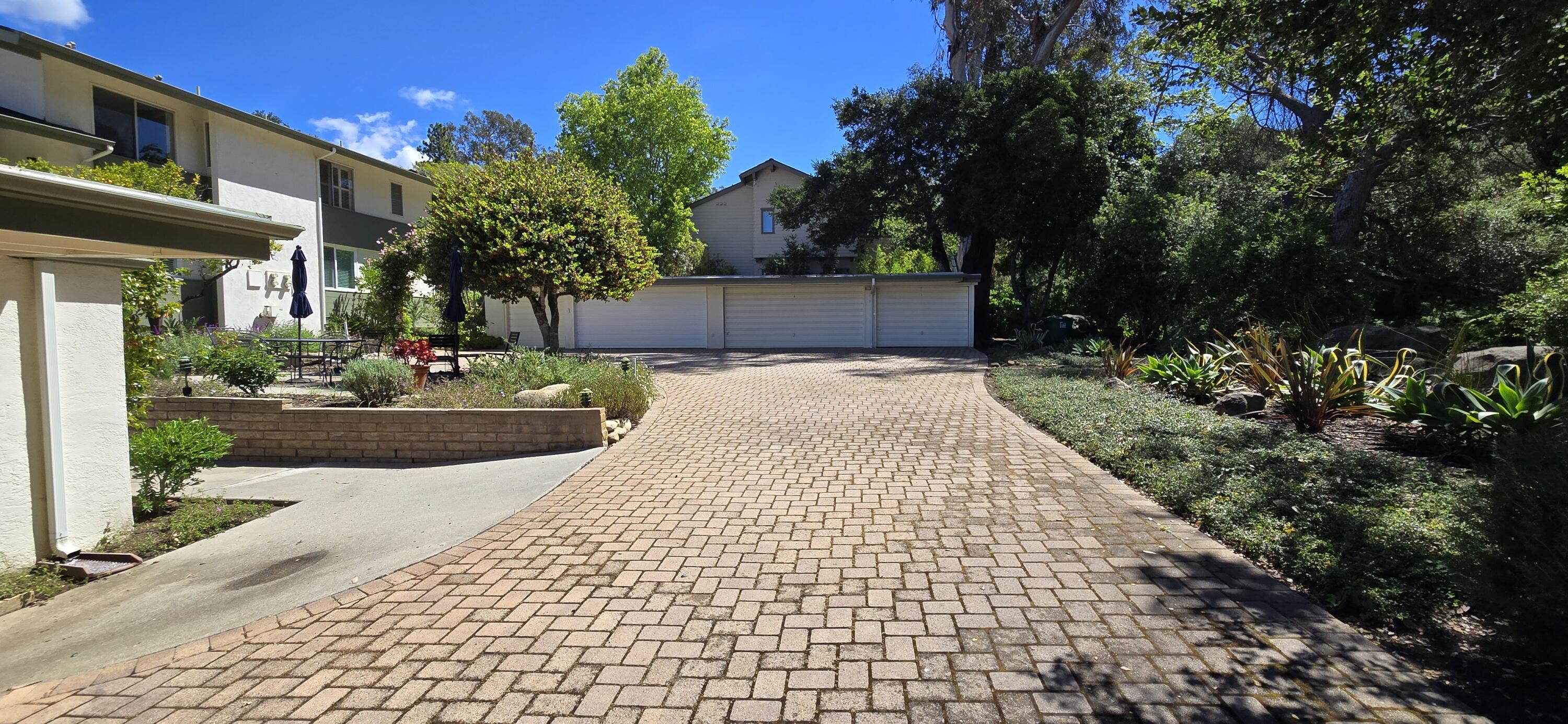 Residential exterior with a wide brick-paver driveway, three garage doors, and mature landscaping.