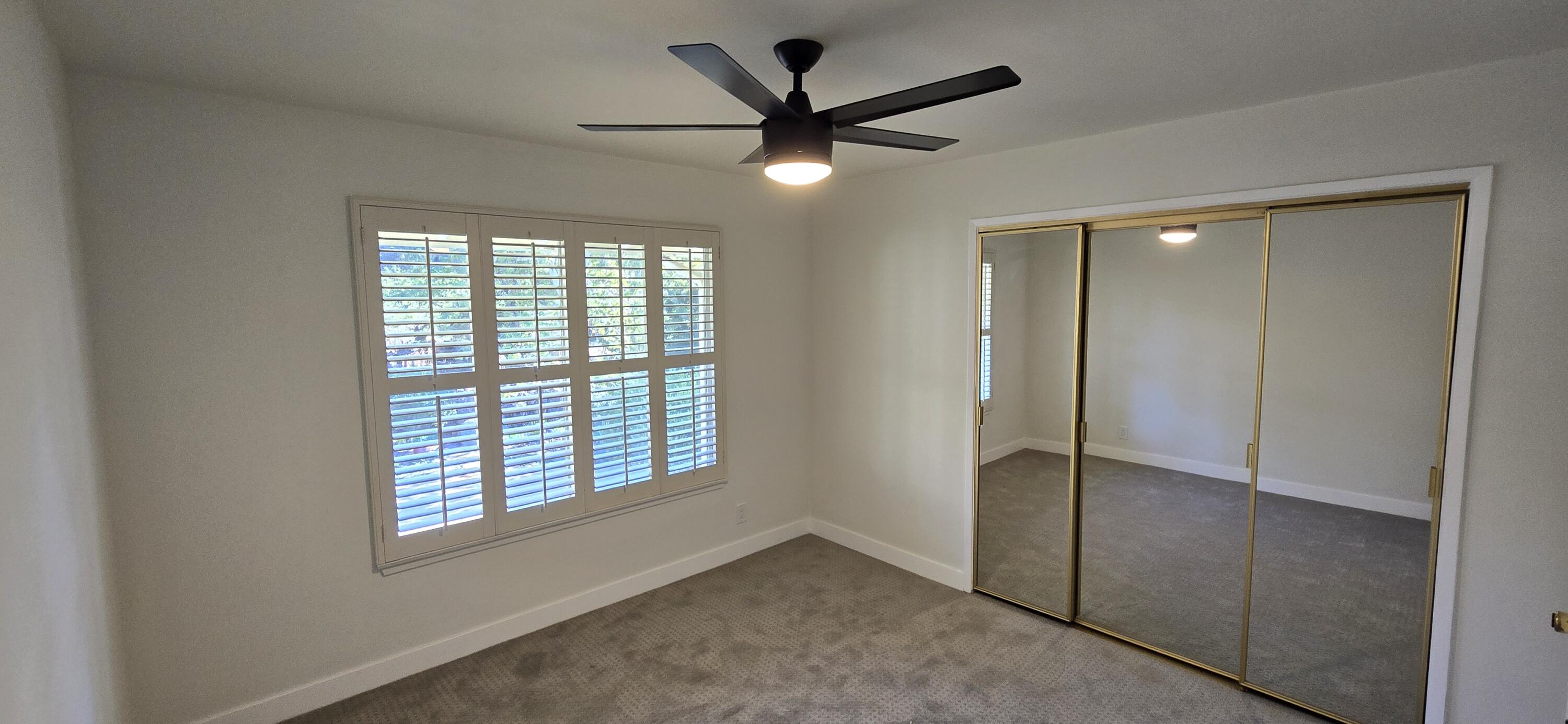 Empty carpeted bedroom with plantation shutters, a ceiling fan, and mirrored sliding closet doors.