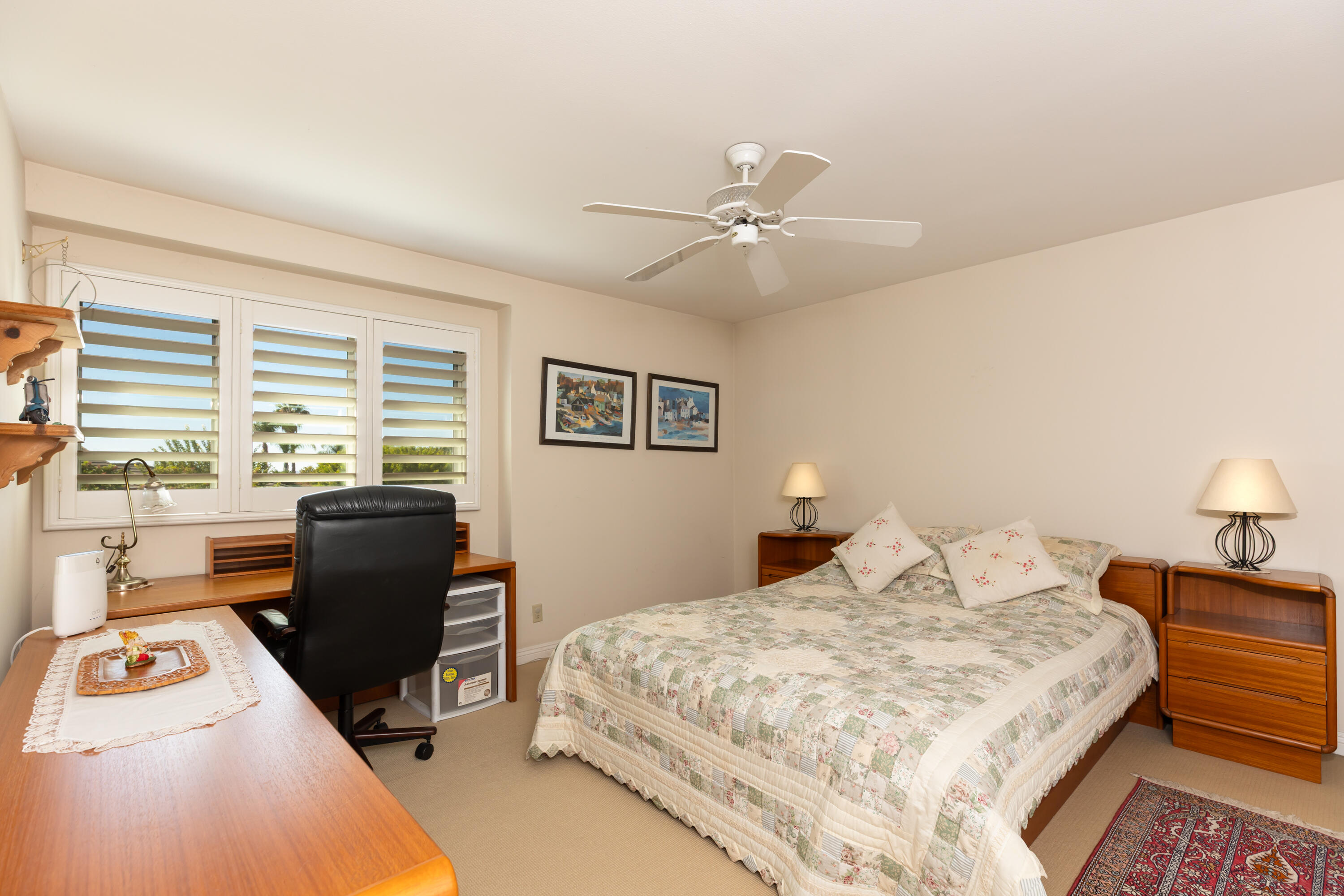 Bedroom with a patterned quilt, wooden furniture, desk, and wide plantation-shuttered windows.