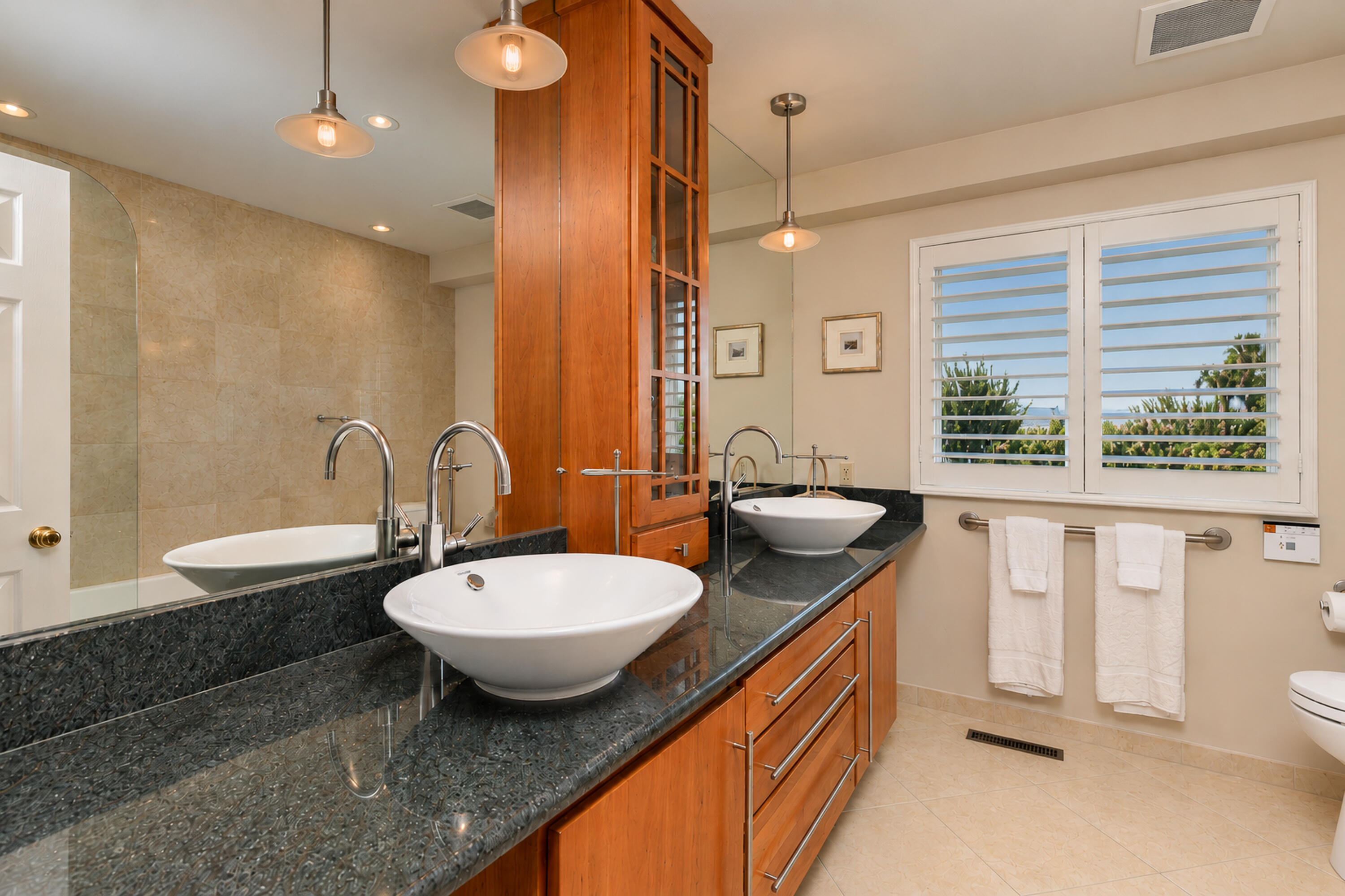 Bathroom with dual vessel sinks, a dark stone vanity, warm wood cabinetry, and plantation-shuttered windows.