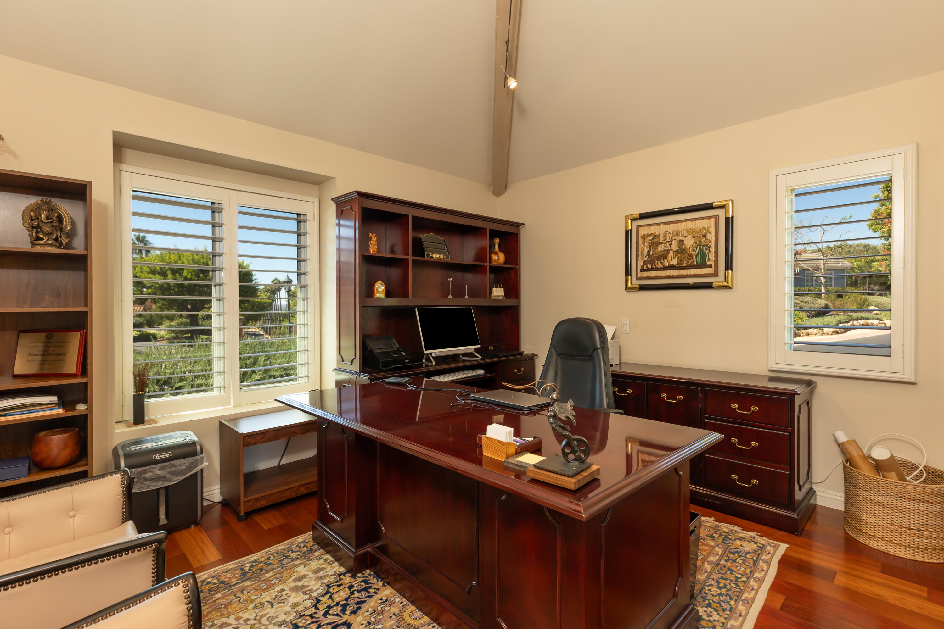 Furnished home office with a polished mahogany desk, built-in shelves, and shuttered windows overlooking greenery.