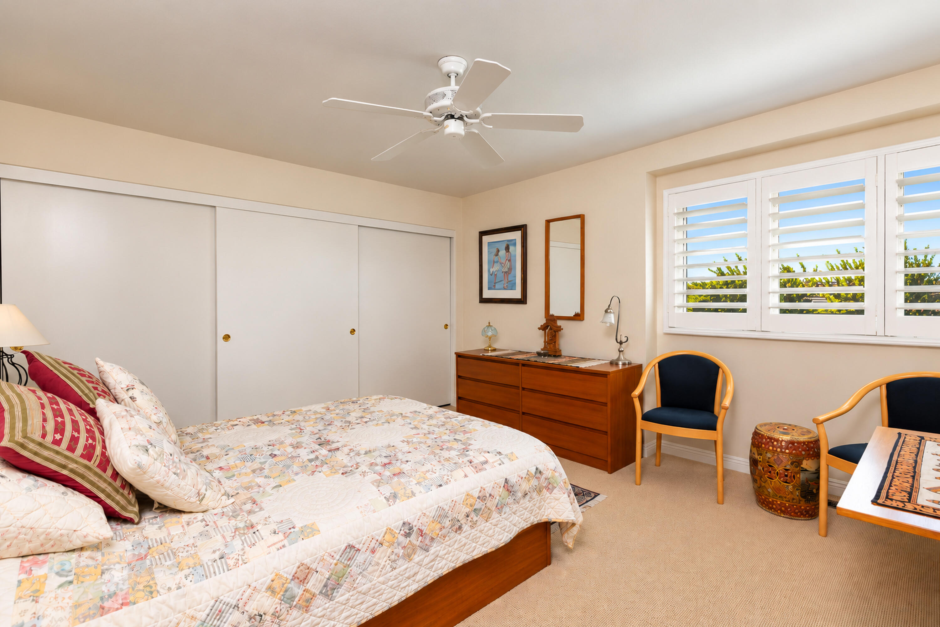 Bedroom with a patchwork quilt, plantation shutters, wooden dresser, and two chairs.