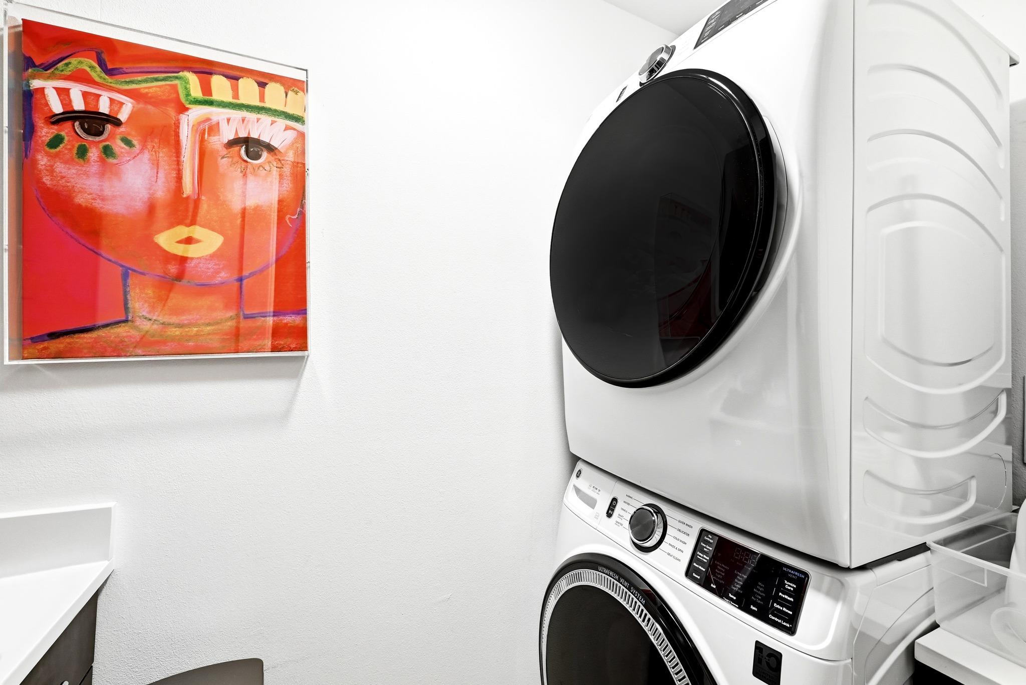 Laundry area with stacked white washer and dryer beside colorful framed portrait artwork.
