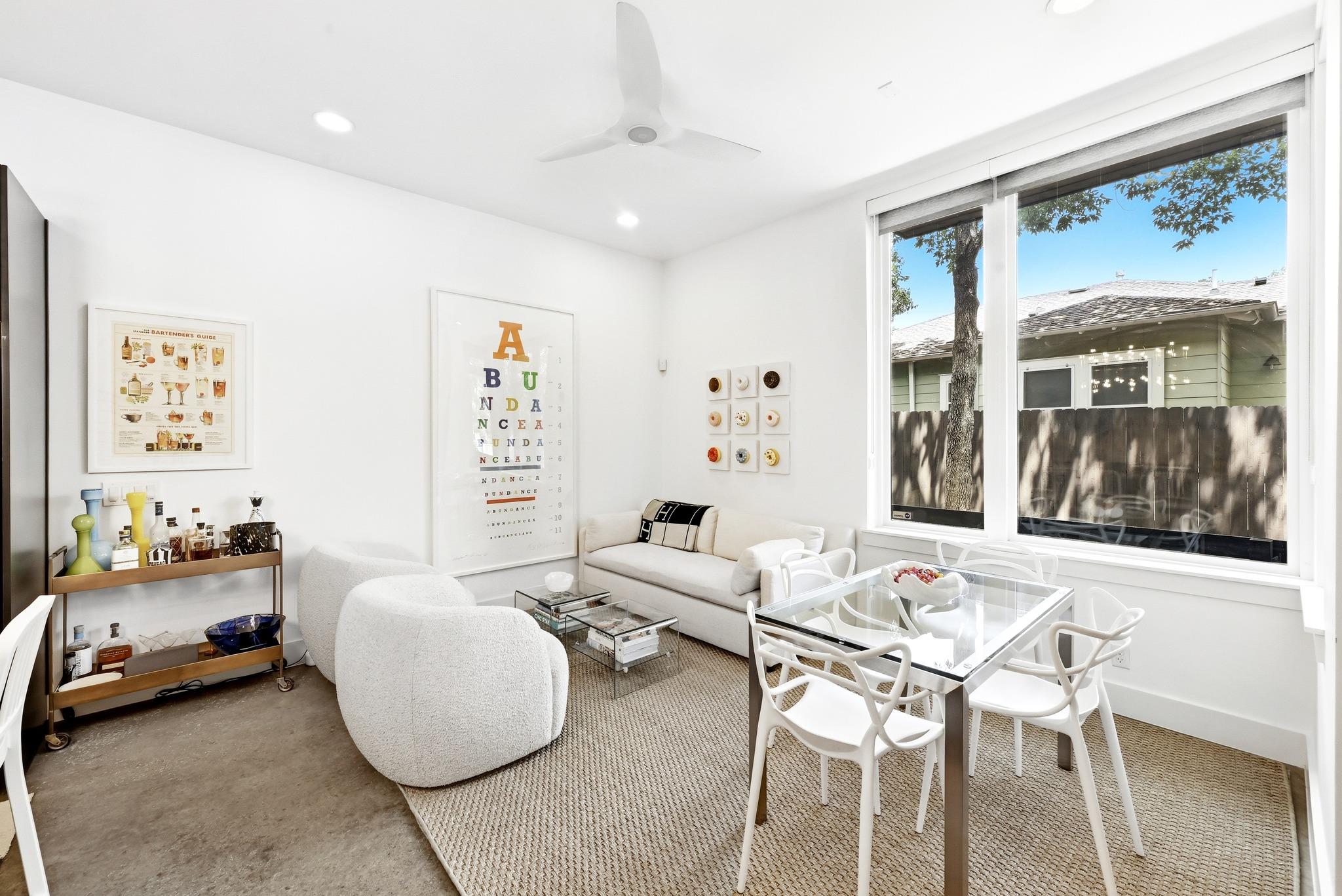 Living and dining room with white seating, a glass table, and large windows overlooking a fenced yard.
