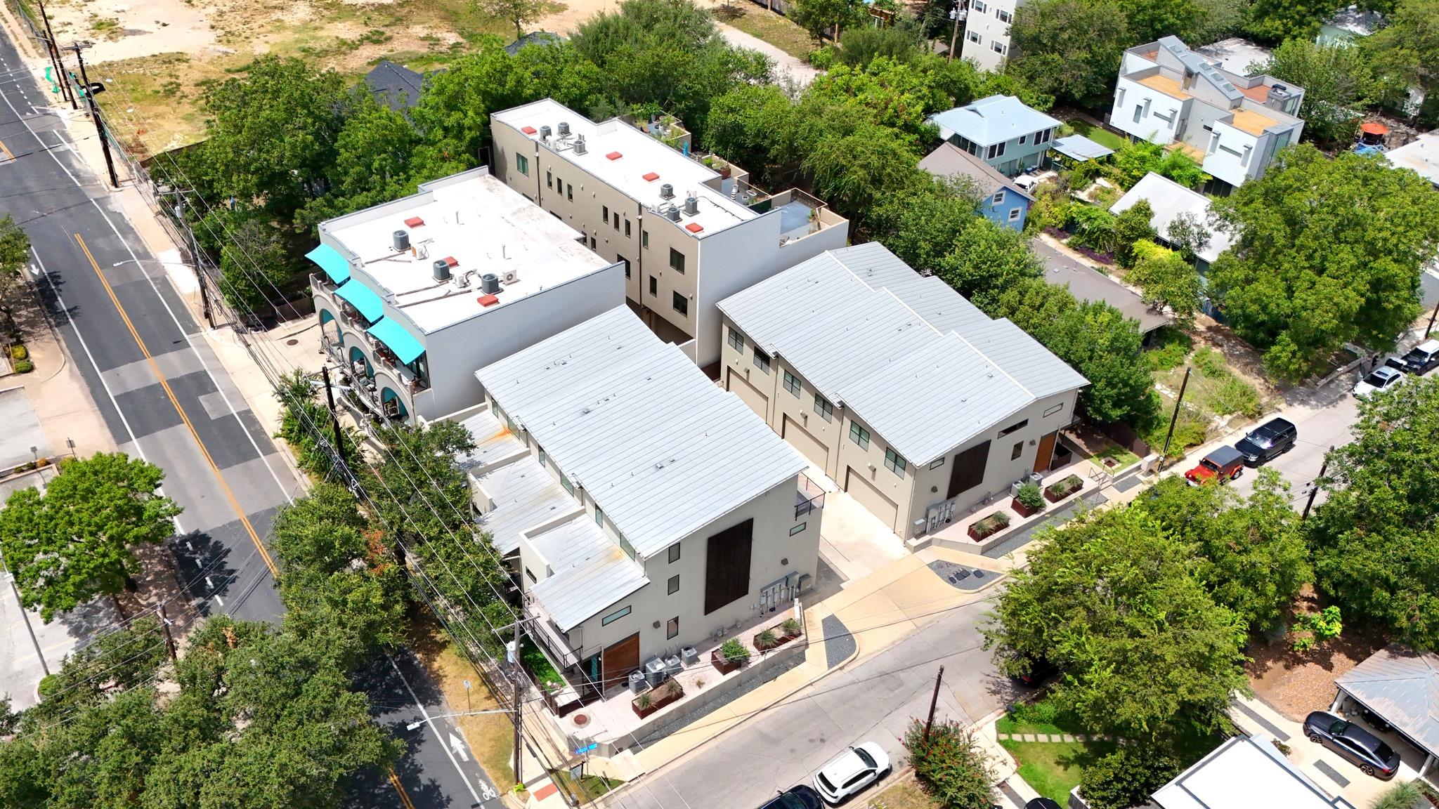 Aerial view of modern multi-unit buildings with white roofs and turquoise awnings beside a tree-lined street.