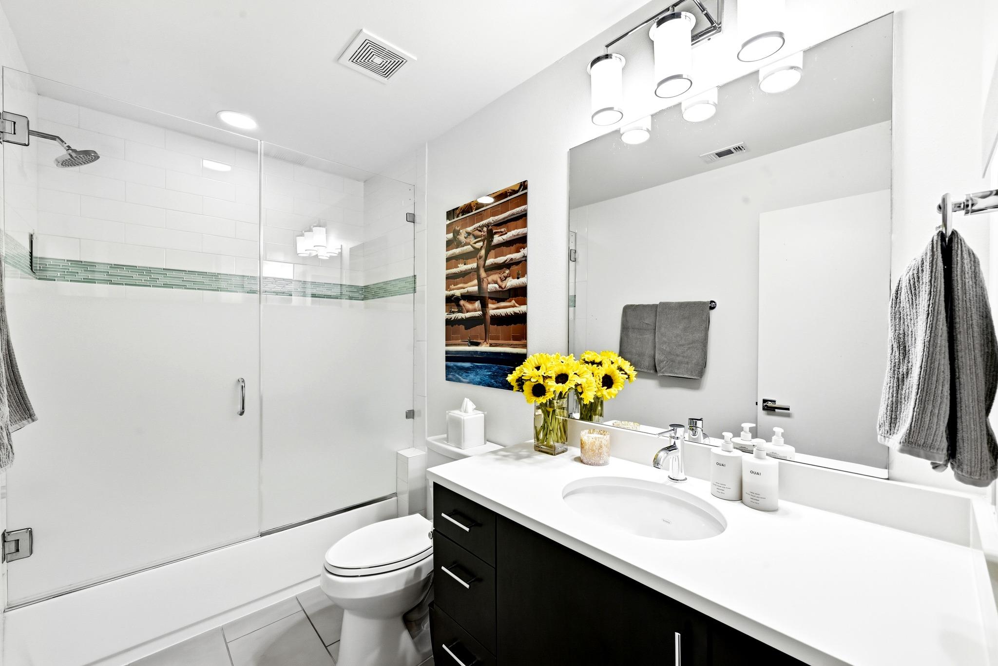 Bright bathroom with a glass shower, green tile accent, dark vanity, and sunflowers on the counter.