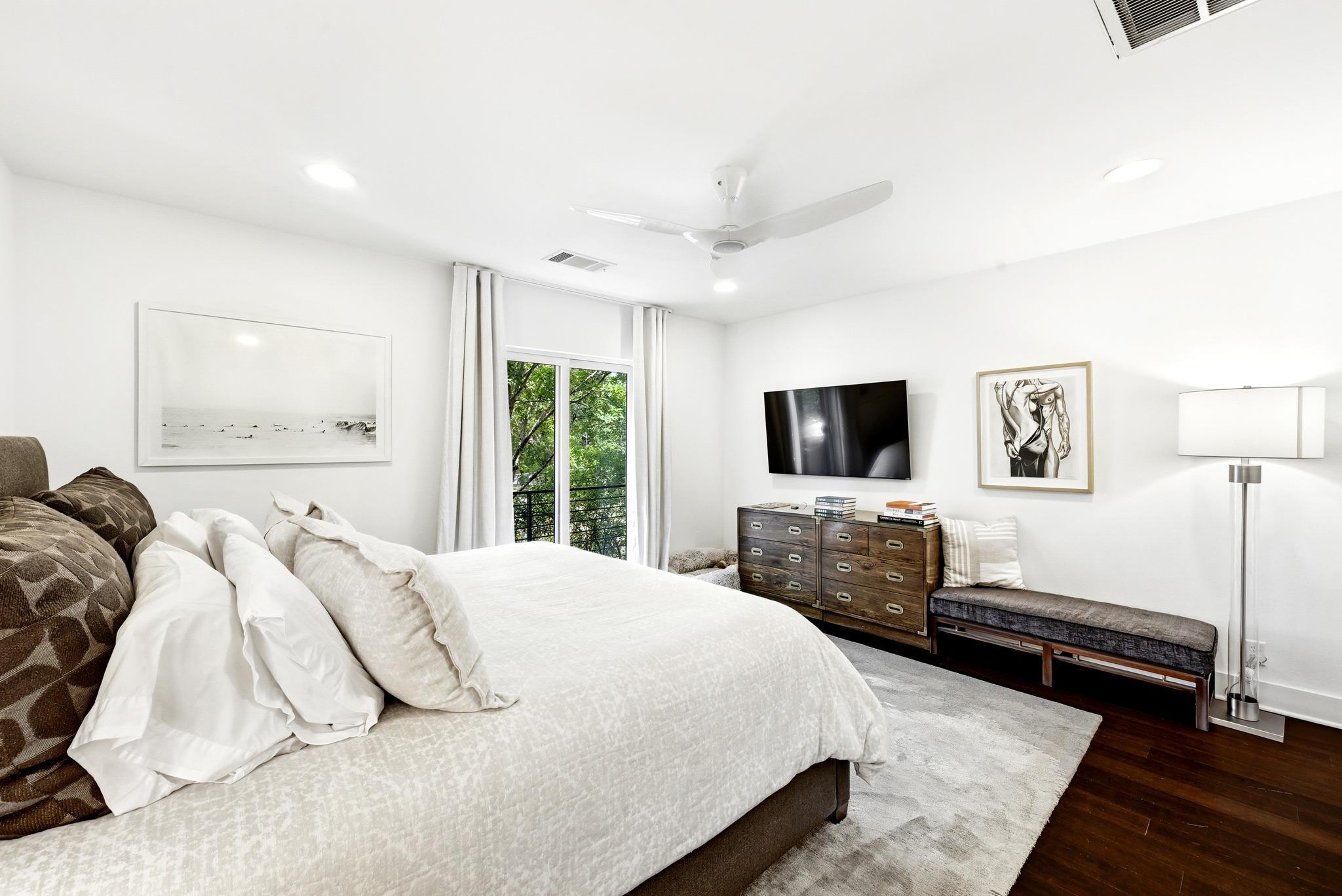 Furnished bedroom with a white bed, dark wood dresser, and leafy views through the window.