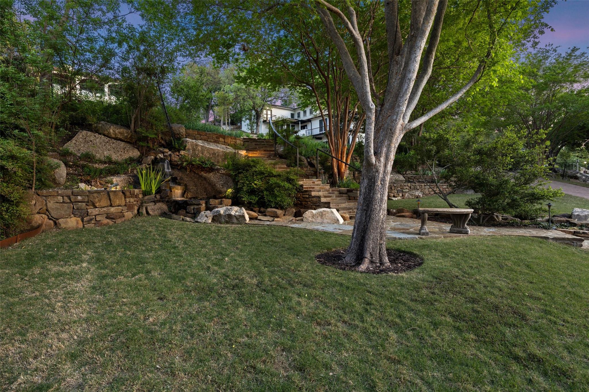 Backyard with a broad lawn, mature trees, stone steps, and a small waterfall beside the house.