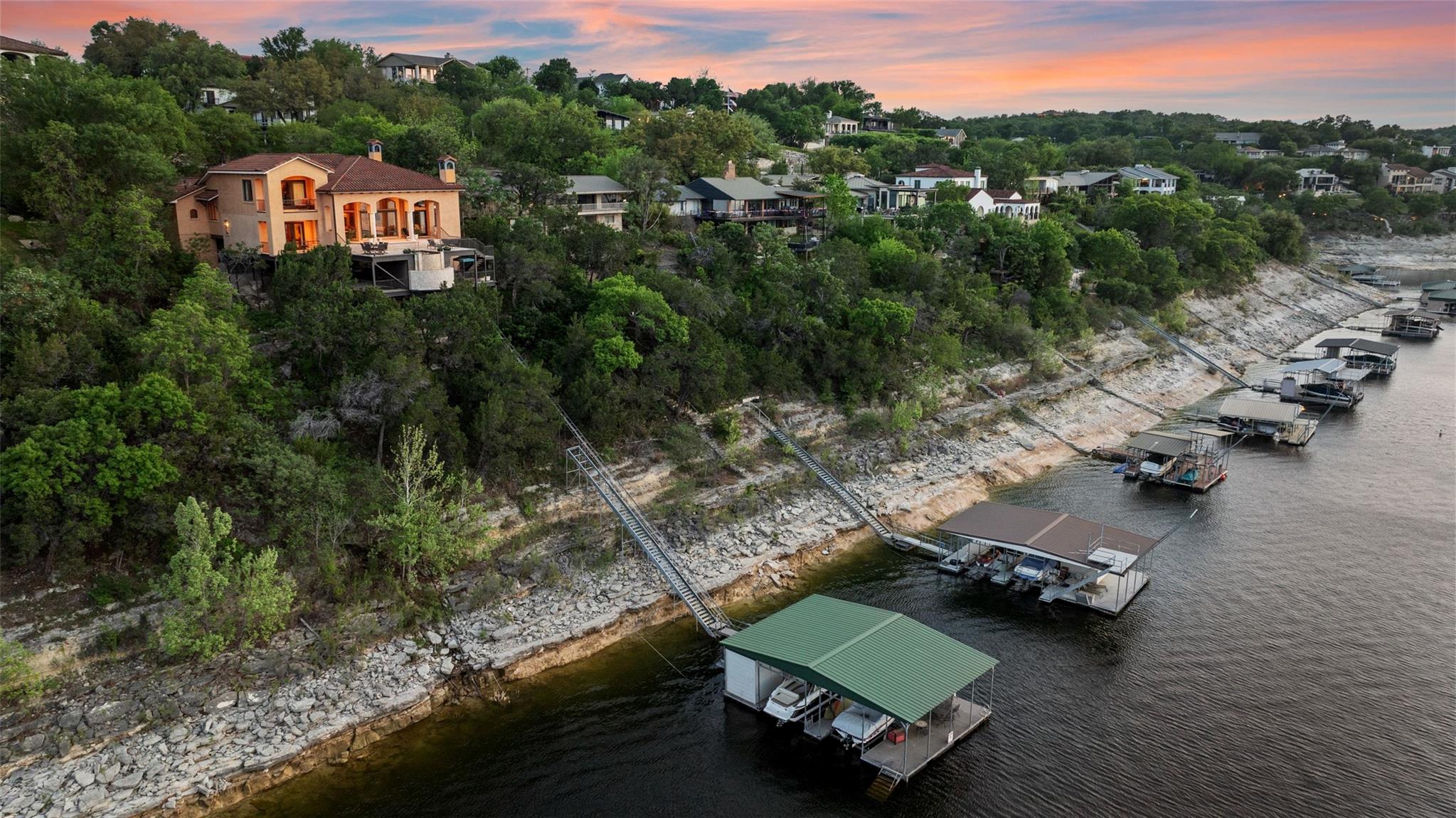 Waterfront homes and boat docks line a rocky lakeshore beneath a pink sunset sky.