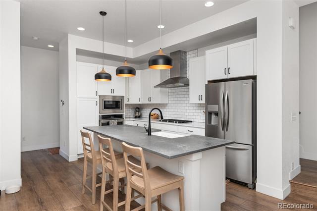 Bright modern kitchen with a large gray island, wooden stools, and stainless steel appliances.