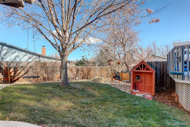 Fenced backyard with a large tree, lawn, red playhouse, and children’s play equipment.
