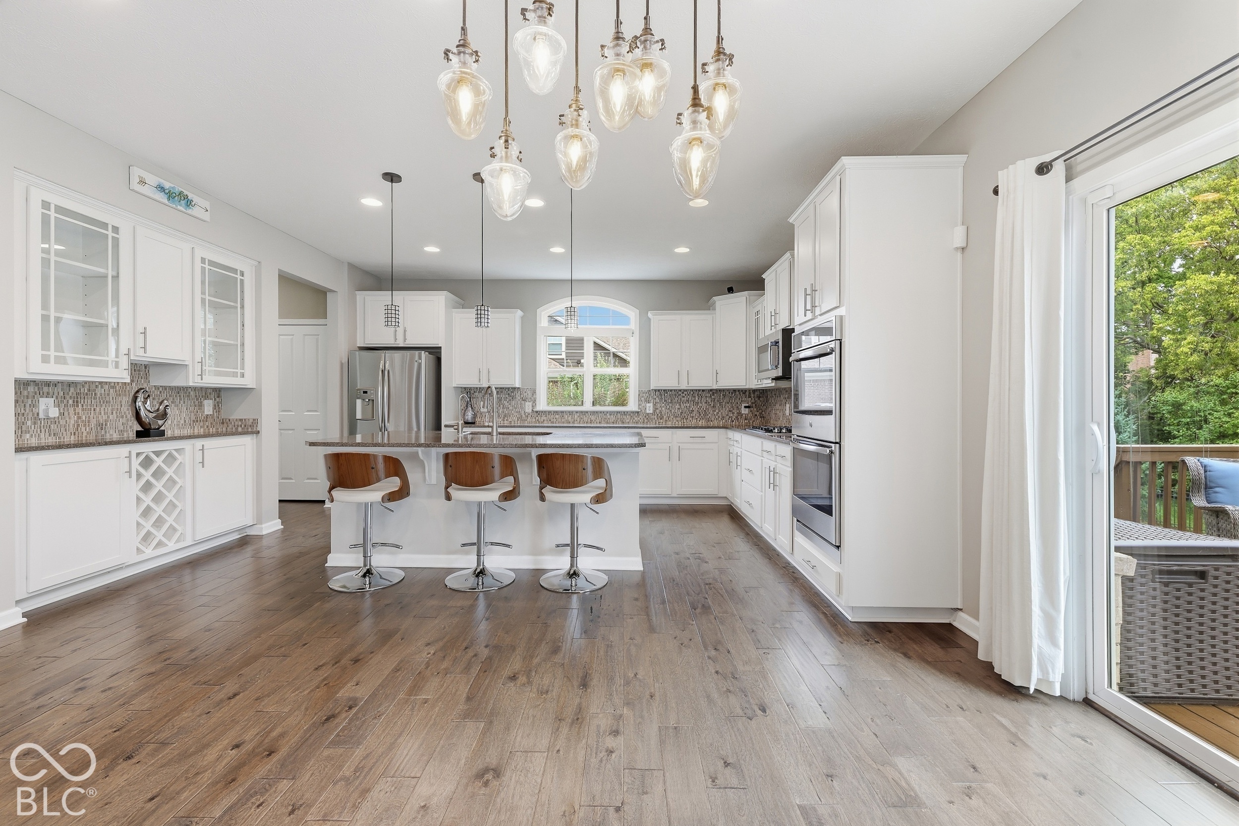 Bright white kitchen with a large island, three bar stools, stainless steel appliances, and sliding glass doors to a deck.