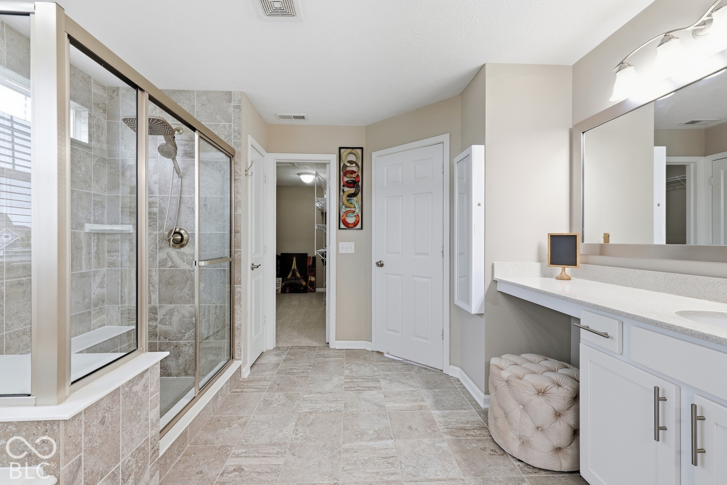 Bathroom with a glass-enclosed tiled shower, white vanity, large mirror, and upholstered stool.