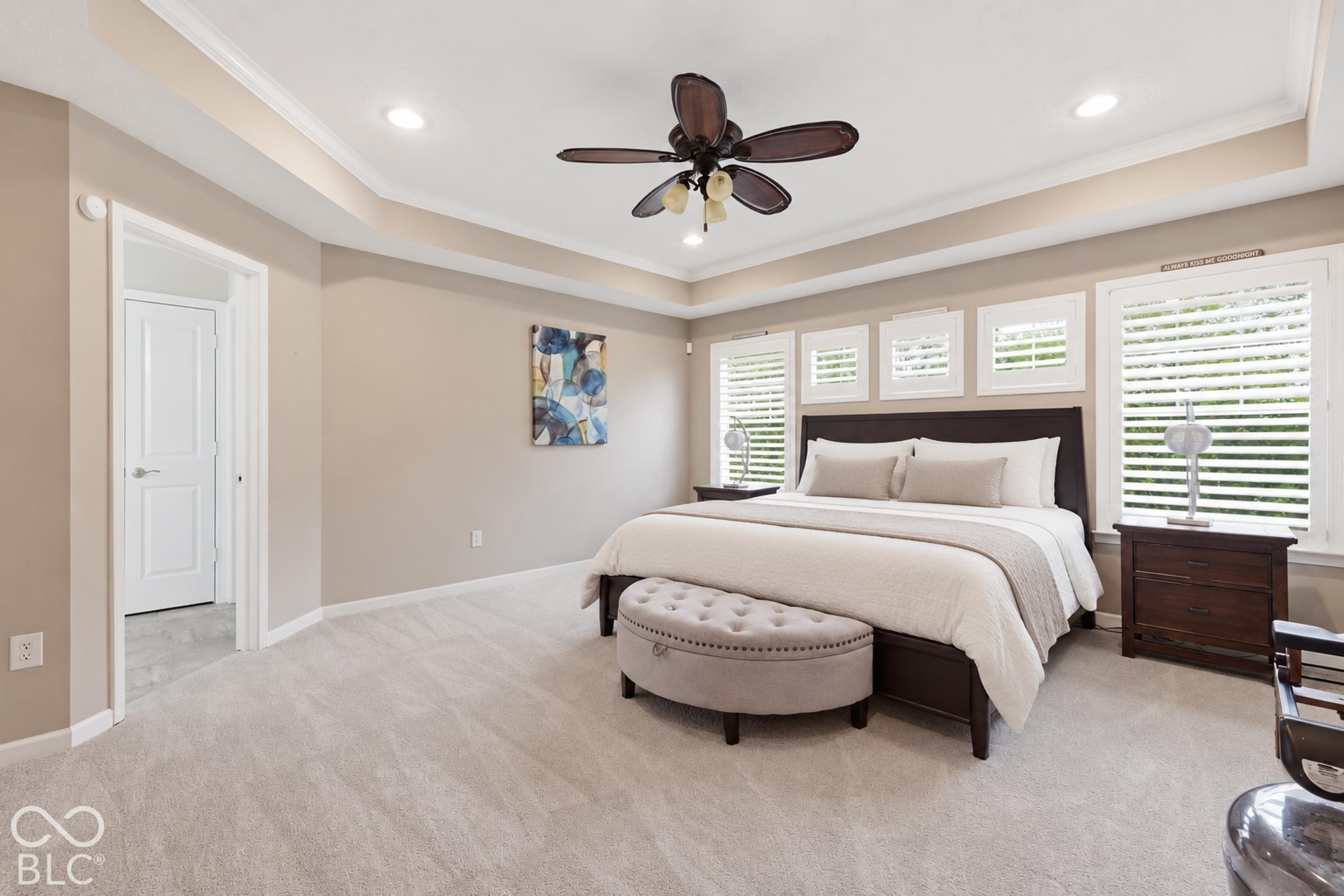 Staged bedroom with a dark wood bed, tufted bench, plantation shutters, and ceiling fan.