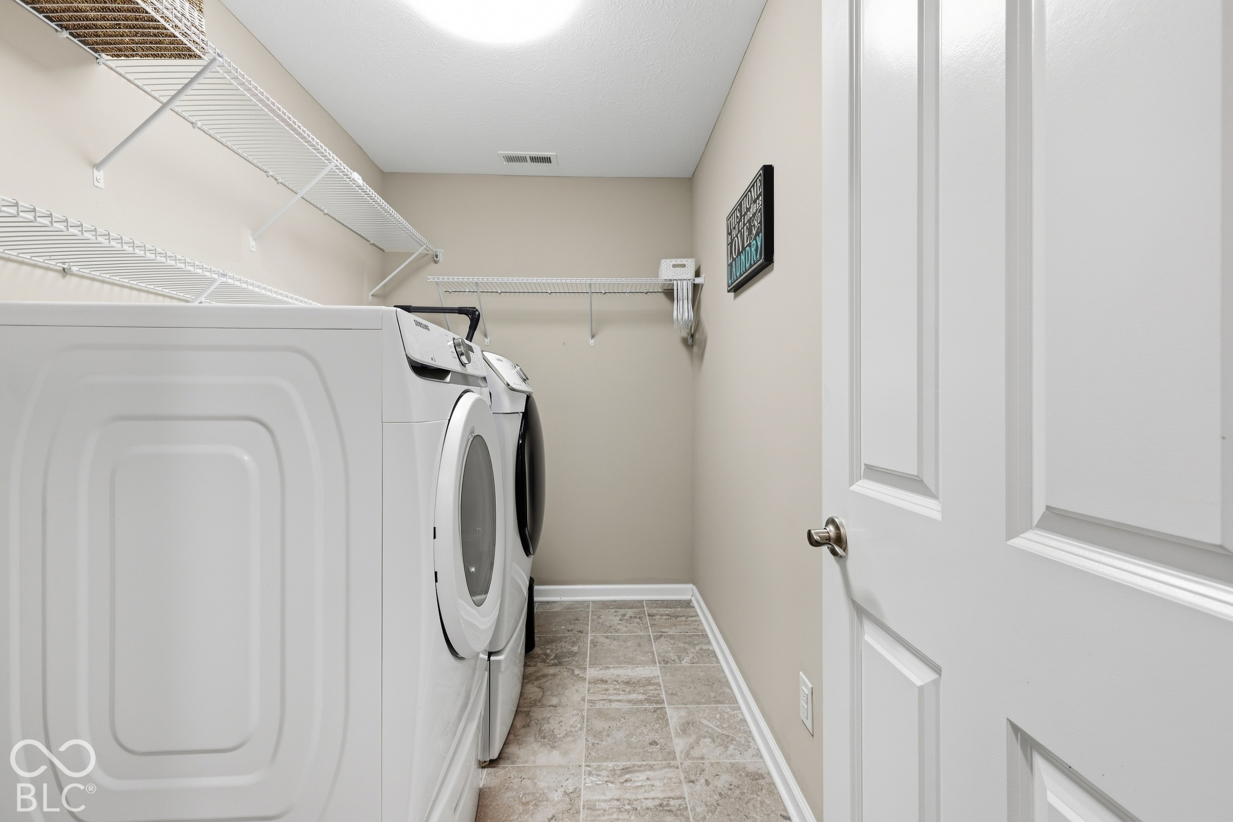 Laundry room with front-loading washer and dryer, wall-mounted wire shelving, and tiled floor.