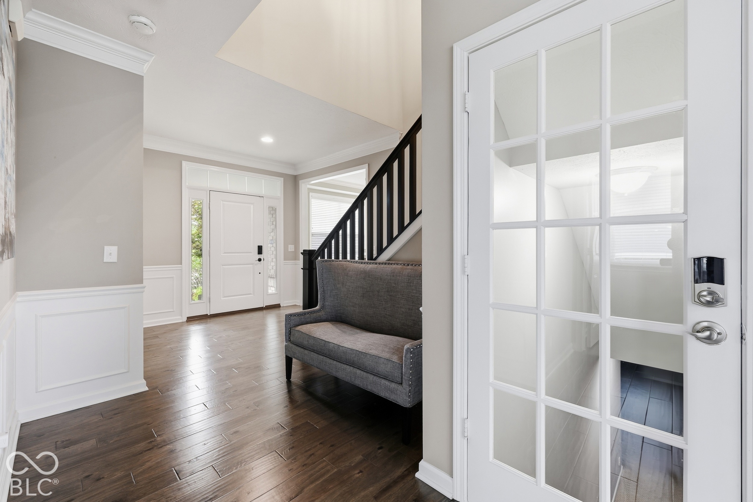 Entry foyer with dark wood floors, a gray bench, and a black staircase railing beside a glass-paneled door.