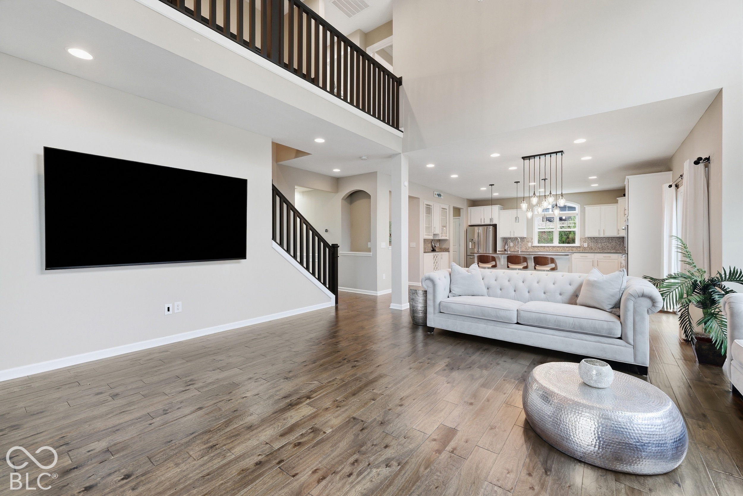 Open-plan living room with hardwood floors, a tufted sofa, dark-railed staircase, and bright white kitchen.